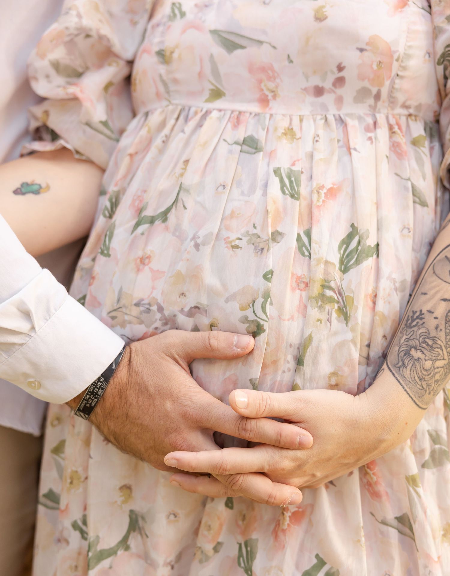 Close-up of couple's hands cradling pregnant belly in floral maternity dress, with visible tattoos on forearm.