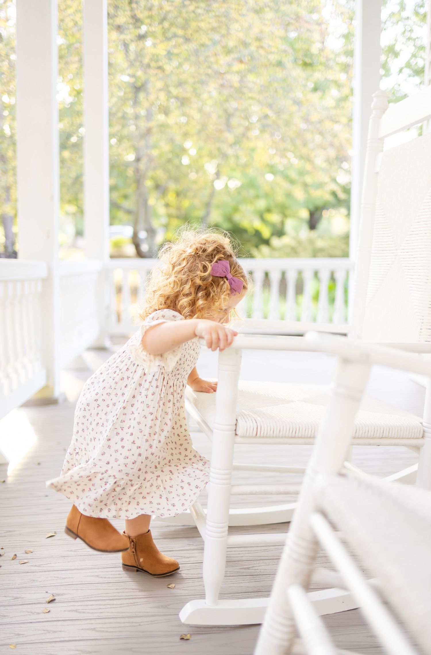 Curly haired toddler in floral dress and brown boots plays on white porch with rocking chairs on sunny day.