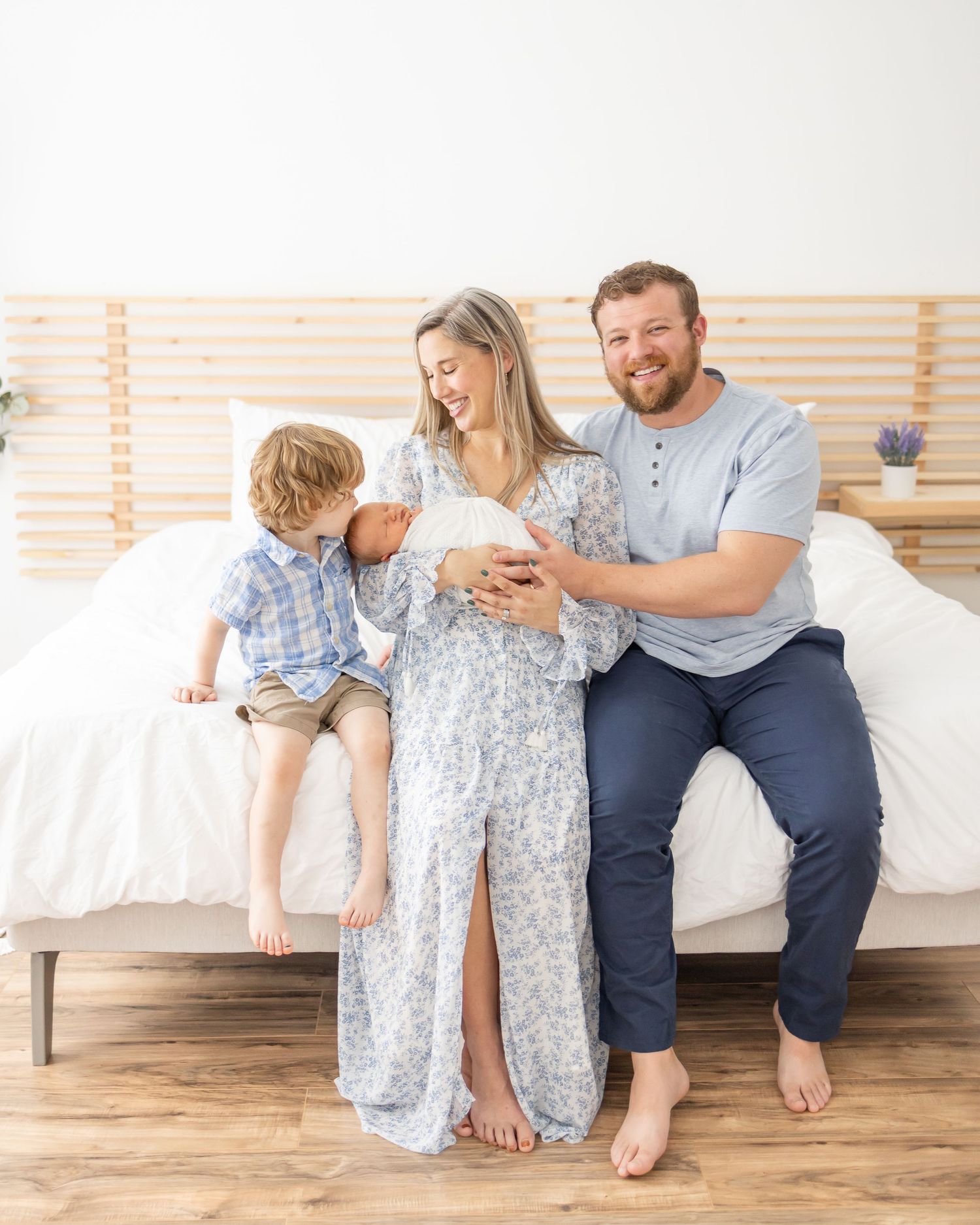 A family sitting on a white bed during their Columbus, GA newborn photography session while their oldest son gives his new brother a kiss.