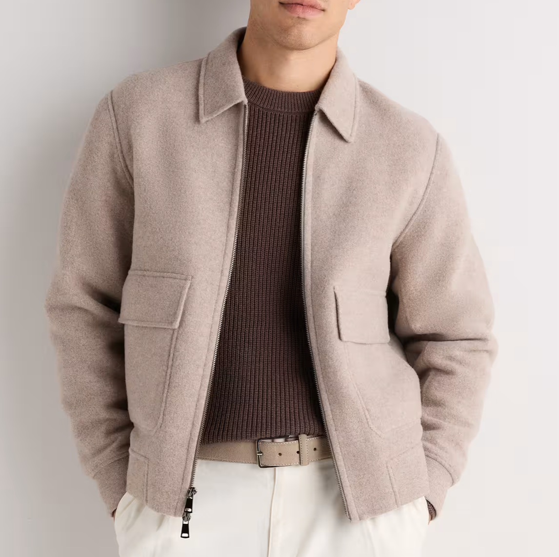 Man wearing a beige wool bomber jacket with patch pockets over a brown ribbed sweater and white trousers.