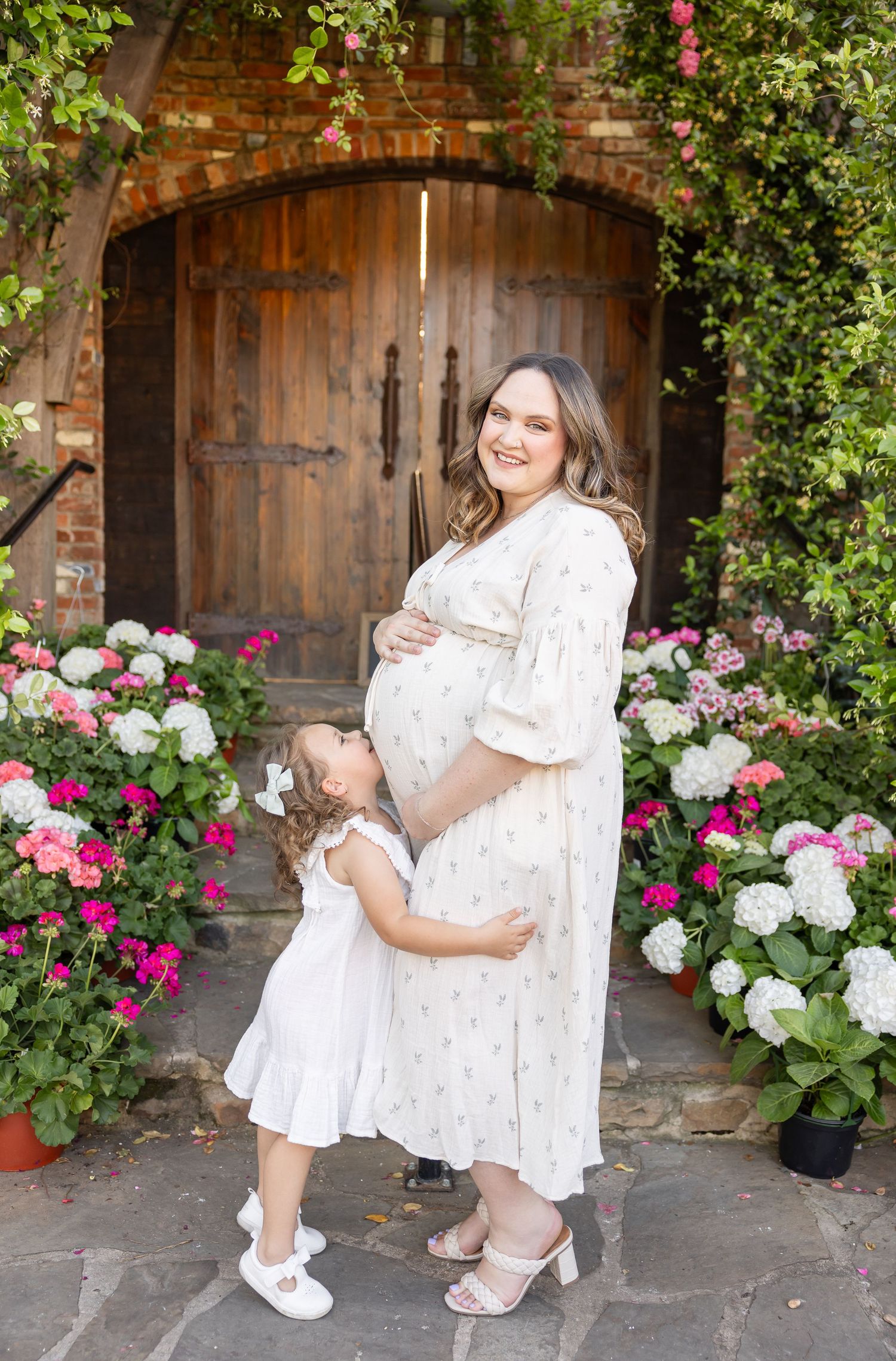 Pregnant woman in floral dress with toddler girl kissing baby bump, standing before wooden door with hydrangeas.
