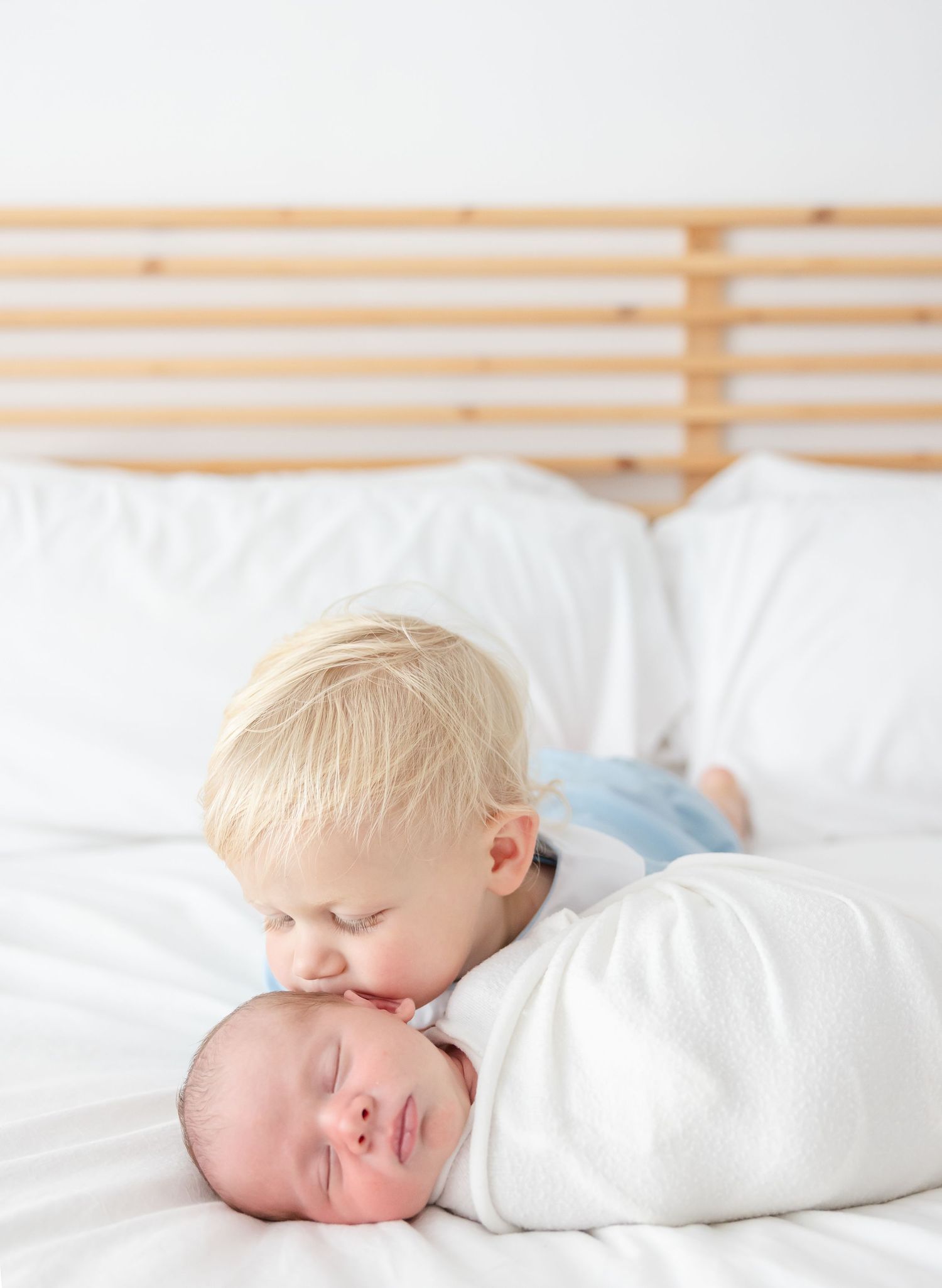 A toddler gently kisses his newborn brother while lying together on white bedding during their newborn photography session in Columbus, GA.