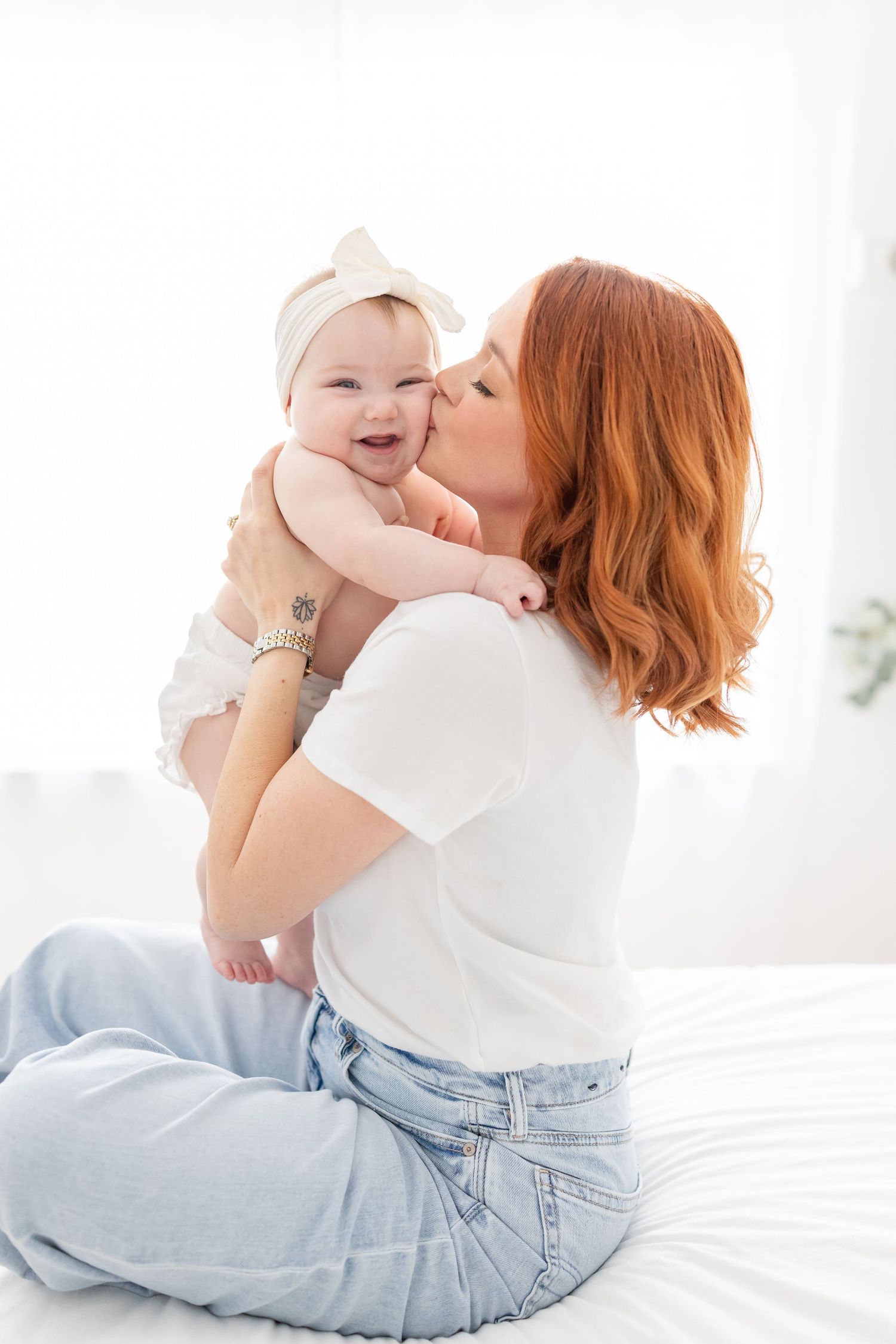 Redheaded mother kissing her smiling baby girl wearing a white bow headband while sitting on a bed.