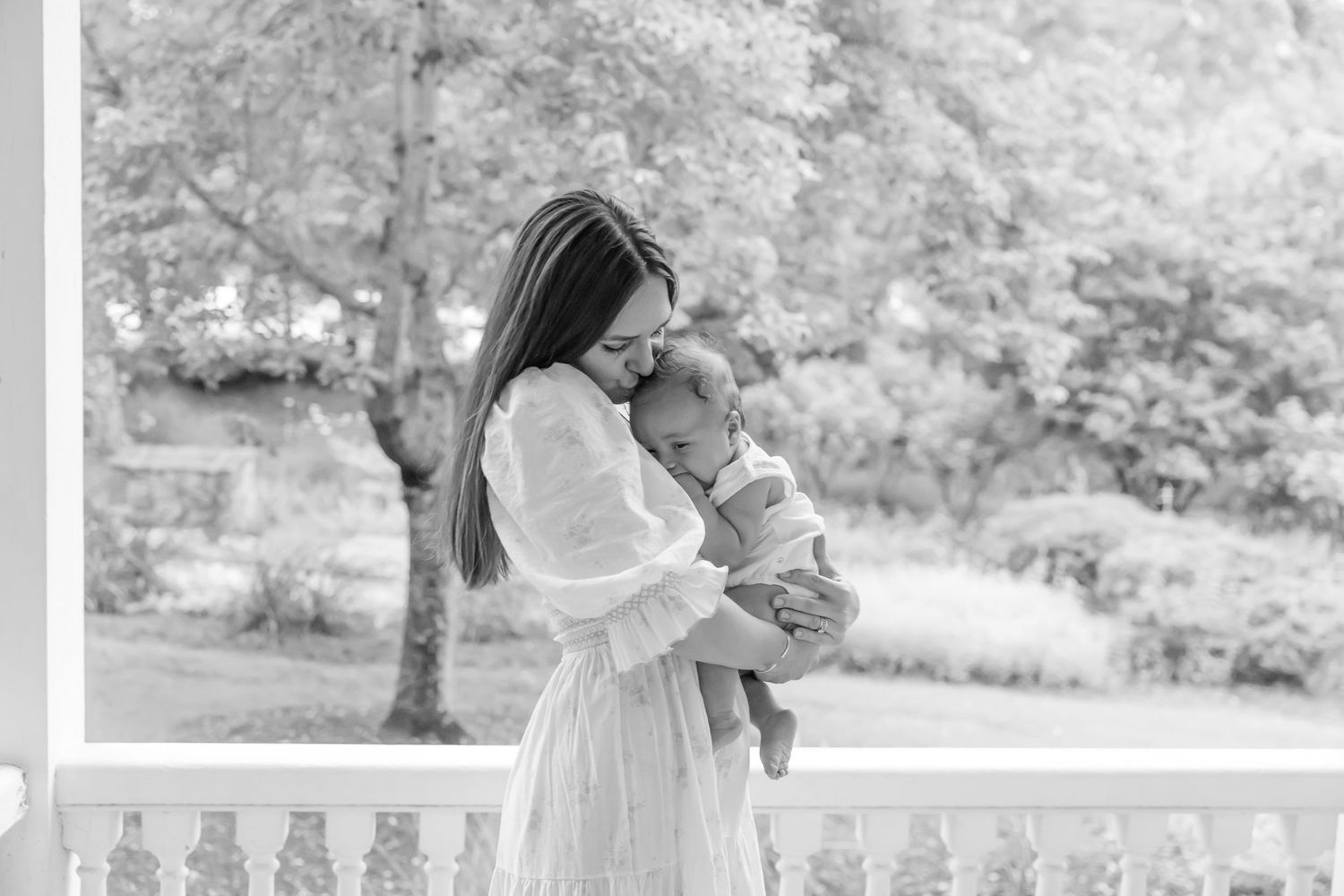 Black and white photo of a mother in a white dress holding her baby on an outdoor porch in Columbus, GA during her family portrait session.