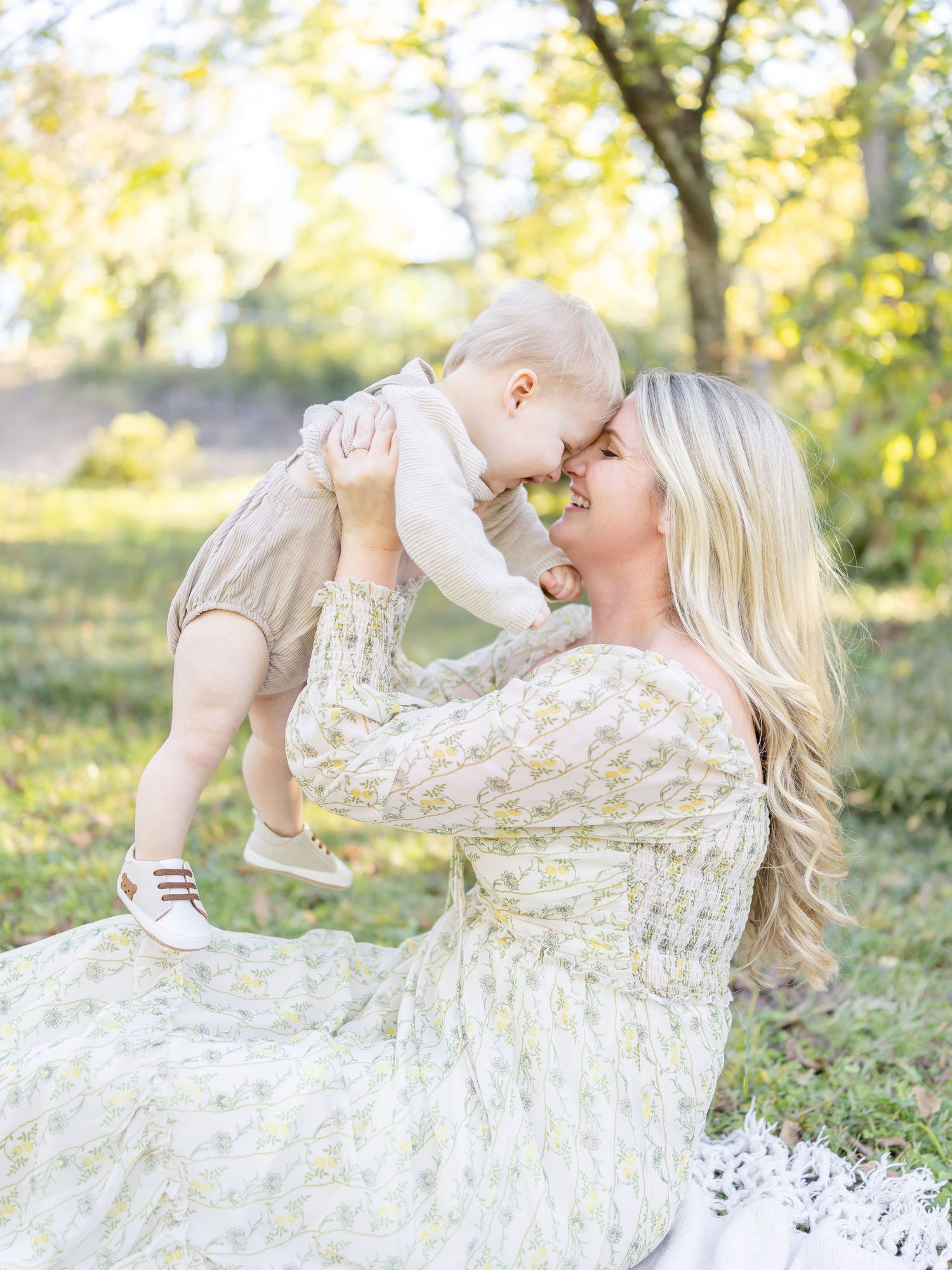 A blonde mother in a floral dress lifts her baby outdoors, sharing a tender nose-to-nose moment in a sunlit park.