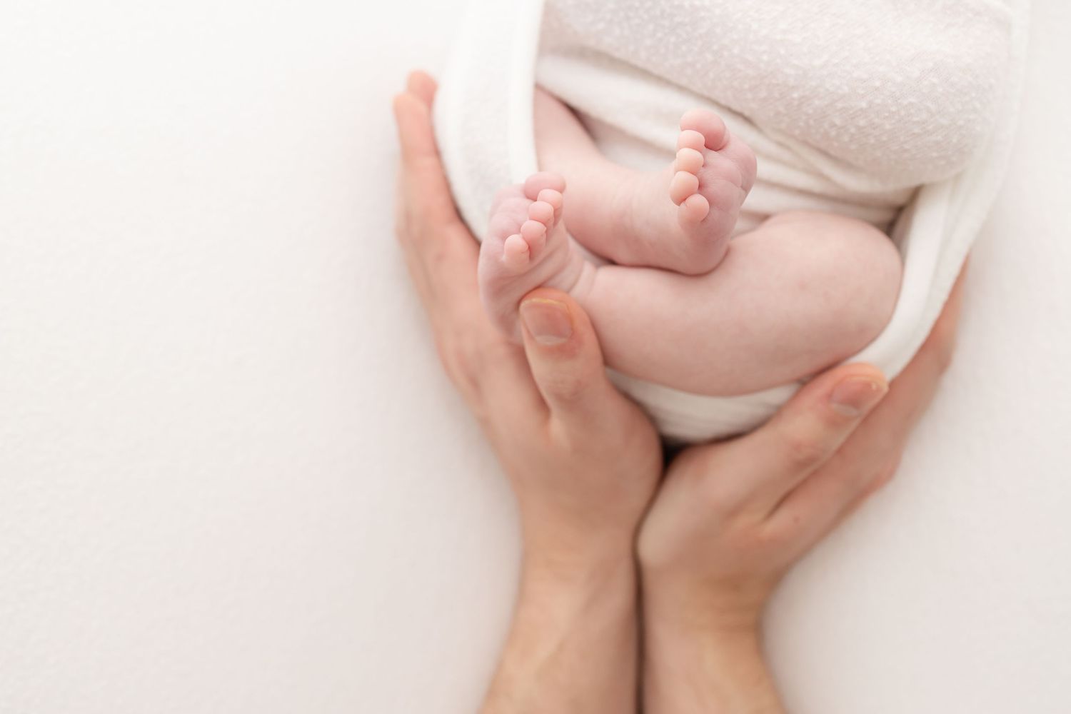 Tiny baby feet being gently cradled in adult hands against a soft white blanket background.