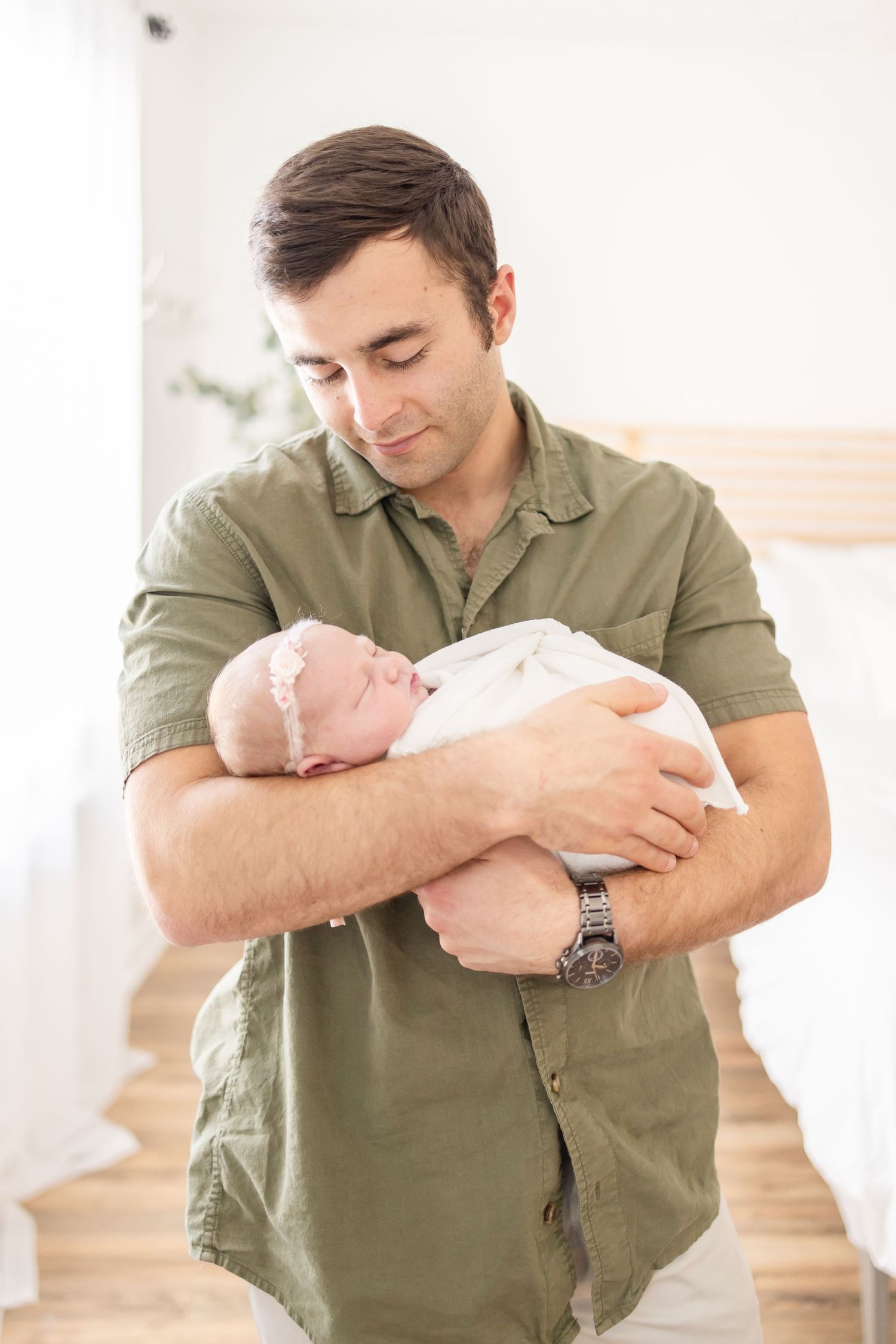 A young father in a green shirt tenderly holds his sleeping newborn baby in a bright, modern bedroom.