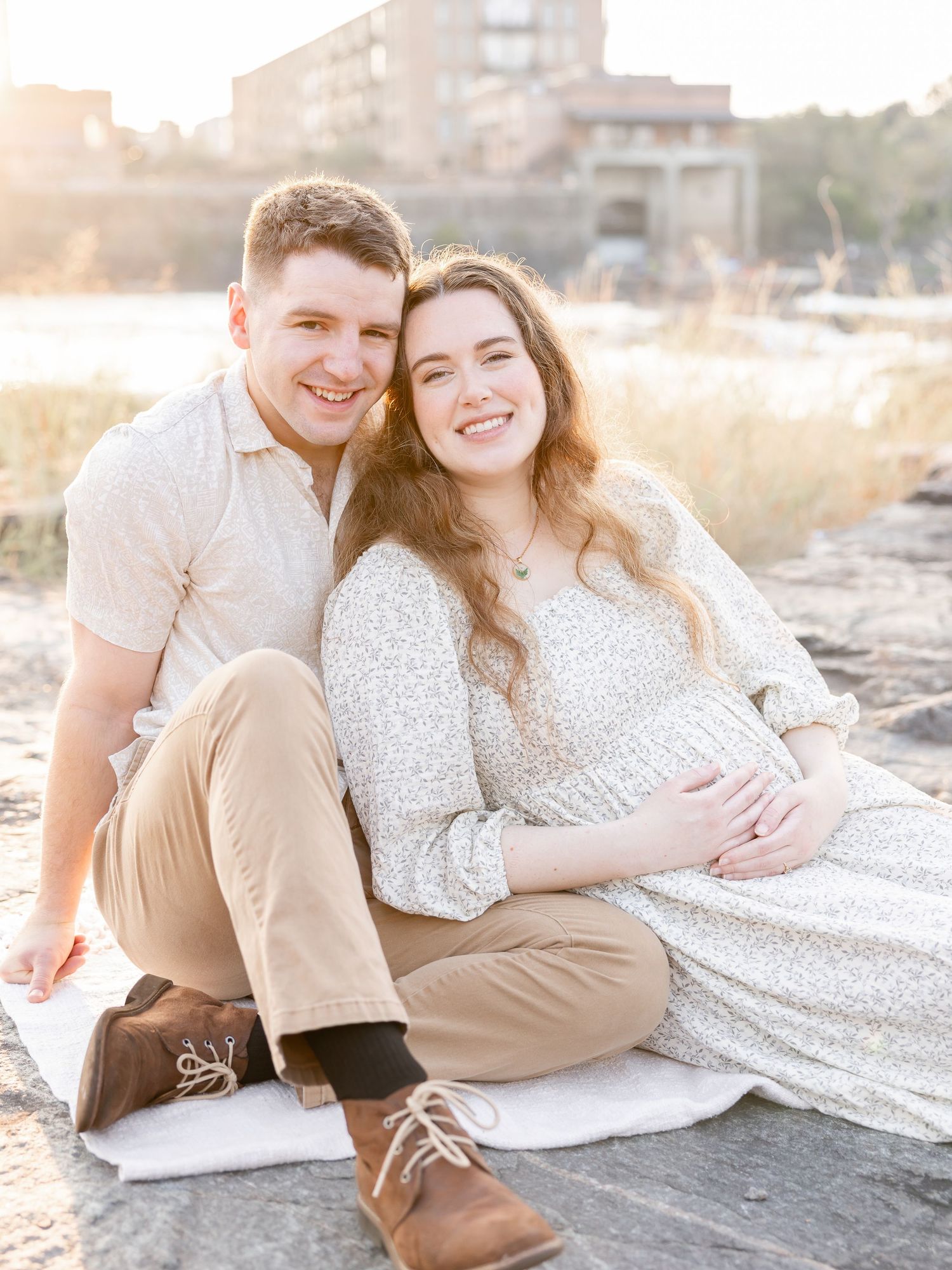 A smiling couple sits on a beach at golden hour, the woman in a floral dress cradling her pregnant belly.