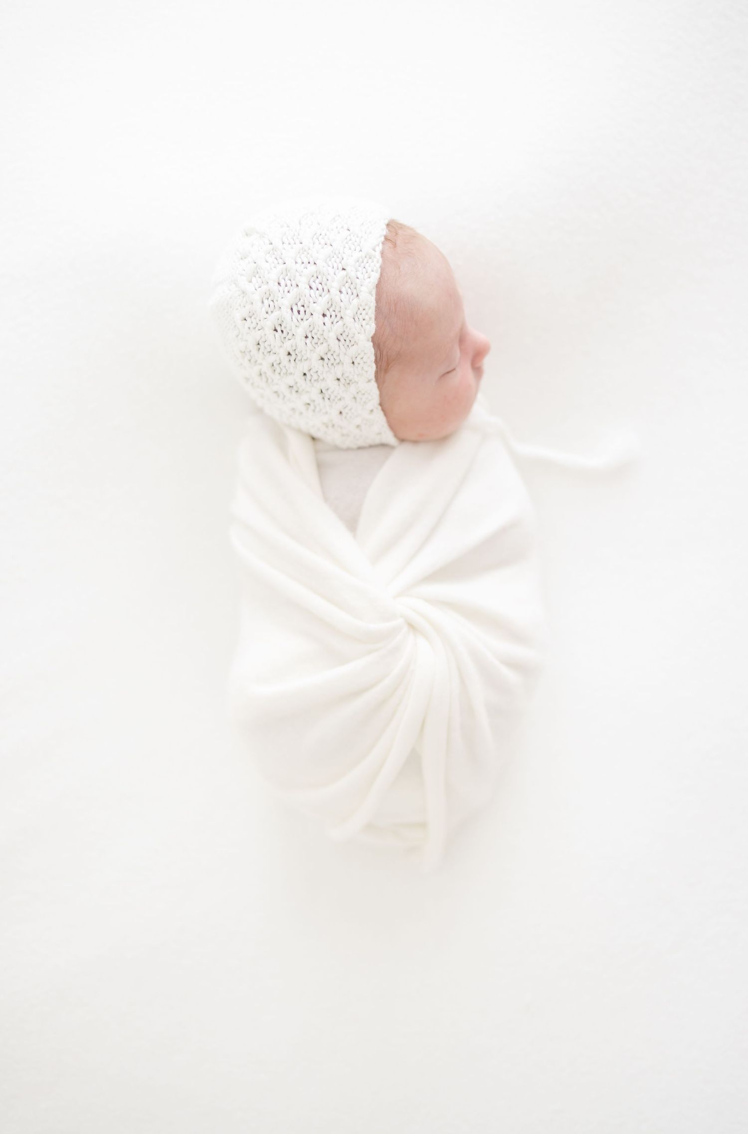 A newborn baby wrapped in white with a knit white bonnet laying on a white background at their Columbus, GA newborn portrait session.