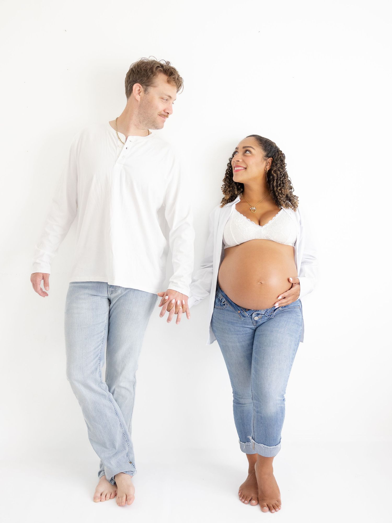 Expecting couple in white tops and jeans holding hands, pregnant woman cradling baby bump, smiling at each other.