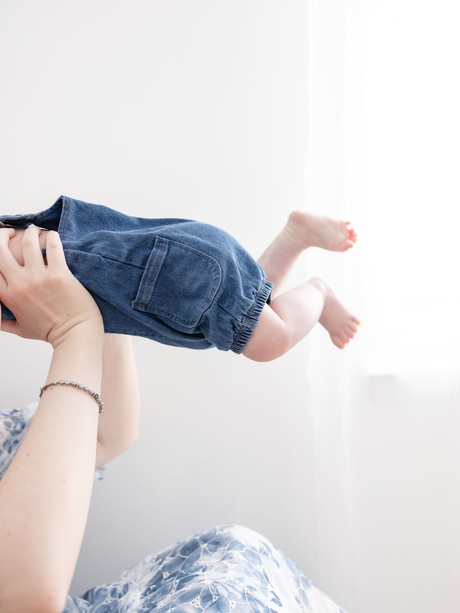 A parent lifting a baby wearing denim overalls in a bright white room, with tiny bare feet visible.
