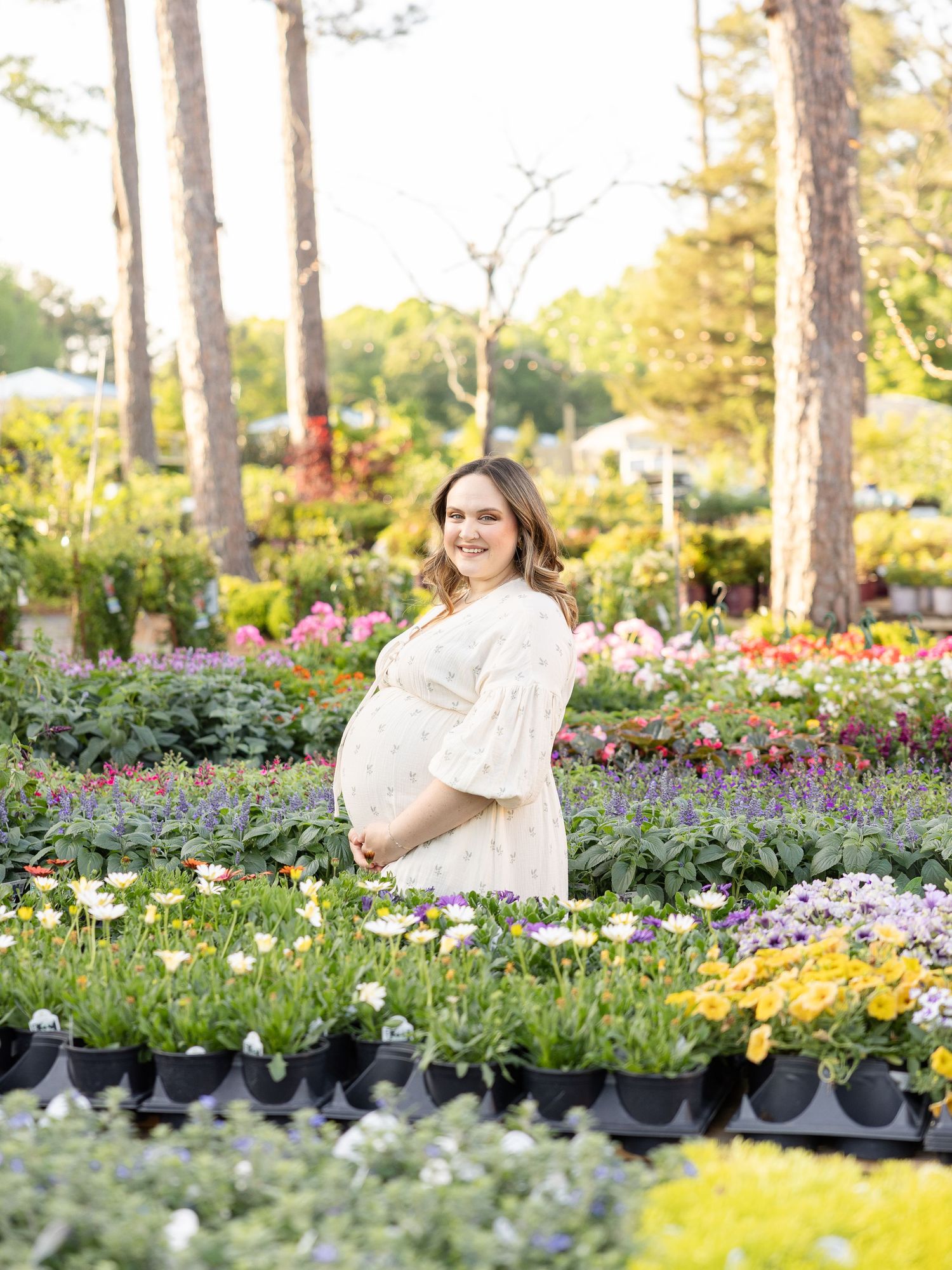 Pregnant mother in white dress smiling among colorful flowers at a garden nursery by Columbus, GA maternity photographer.