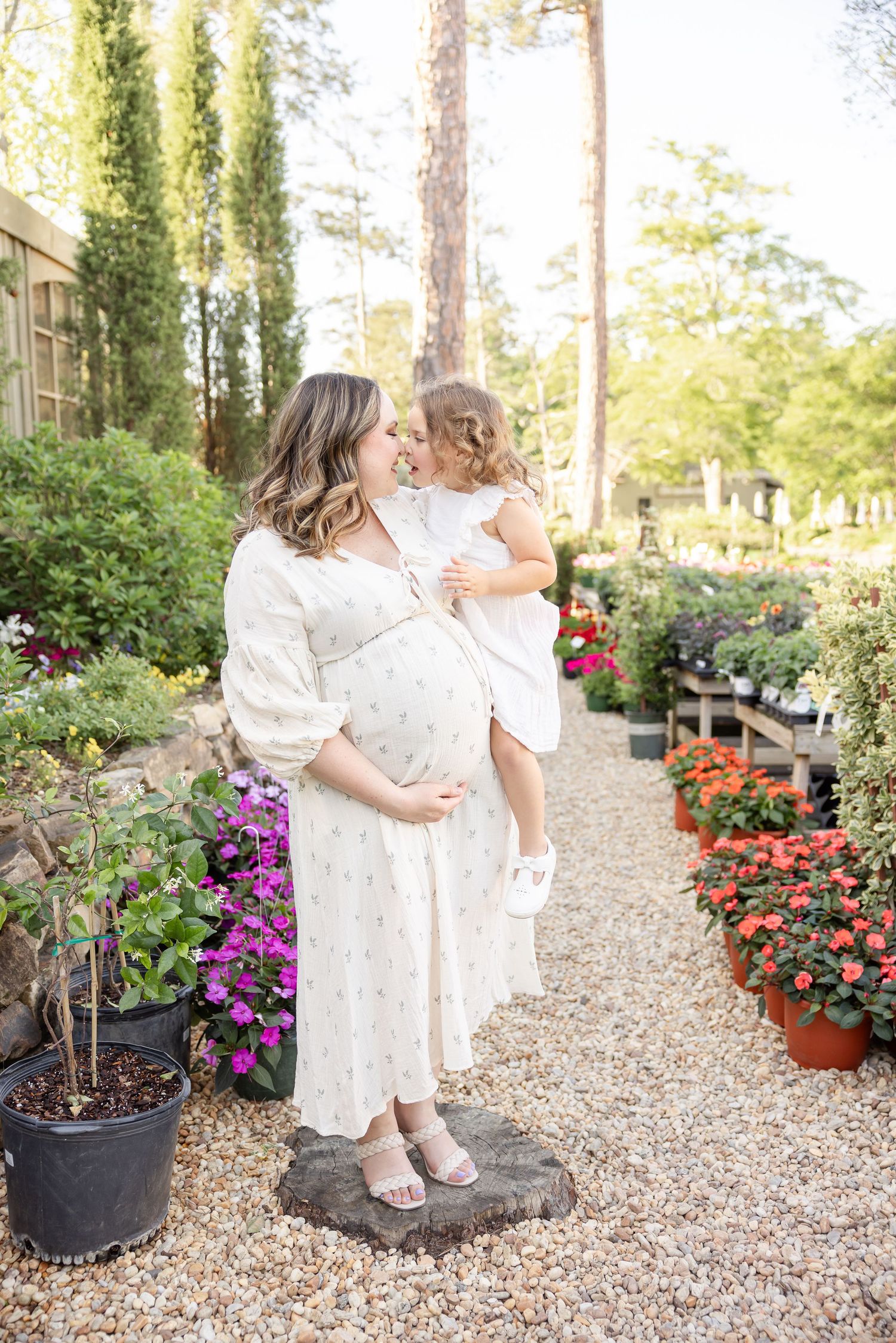 Pregnant woman in floral dress holds her toddler among colorful garden flowers on a sunny day during her maternity portraits in Opelika, AL
