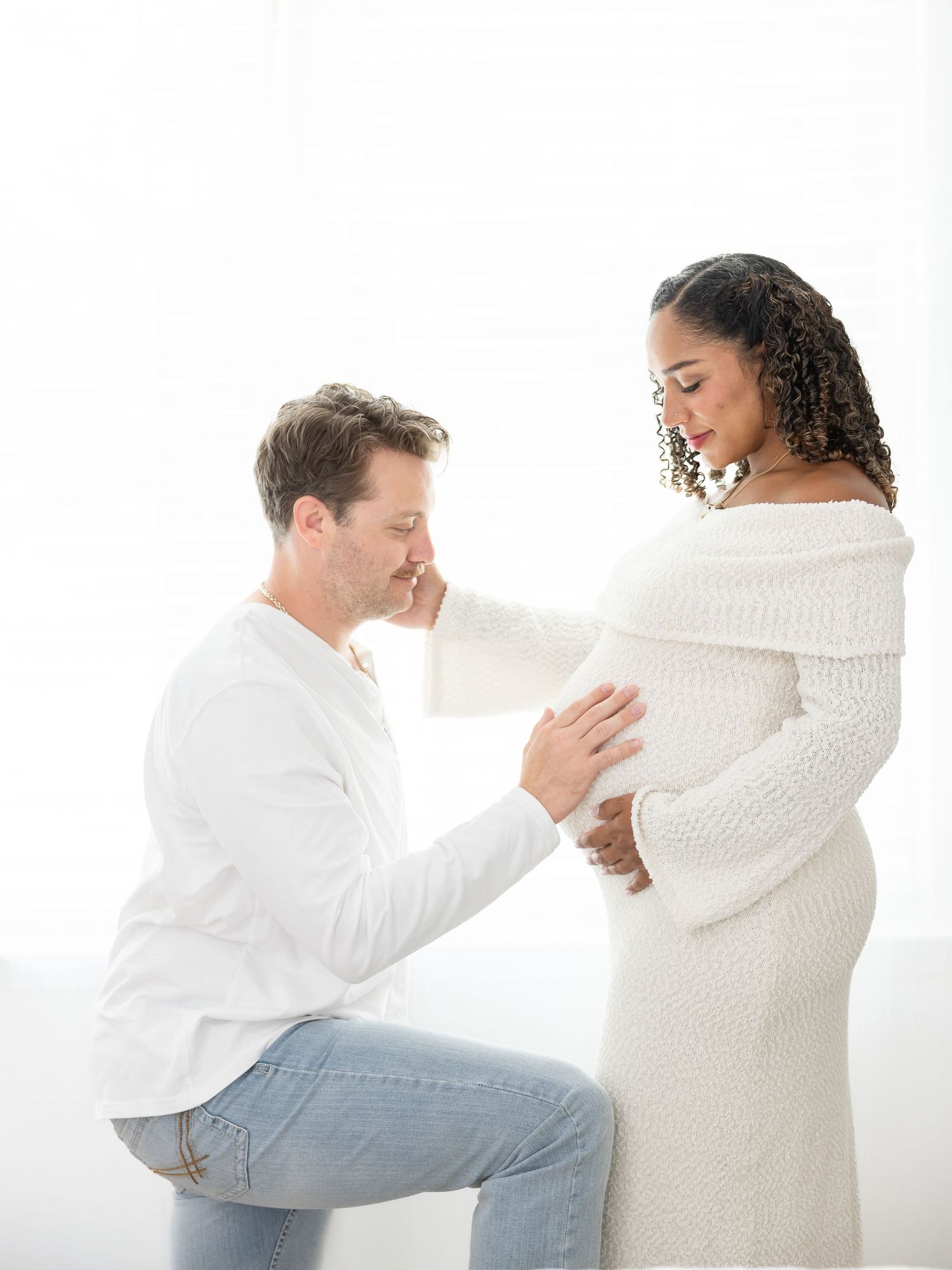A man kneels touching a pregnant woman's belly in a bright white studio maternity photo shoot.