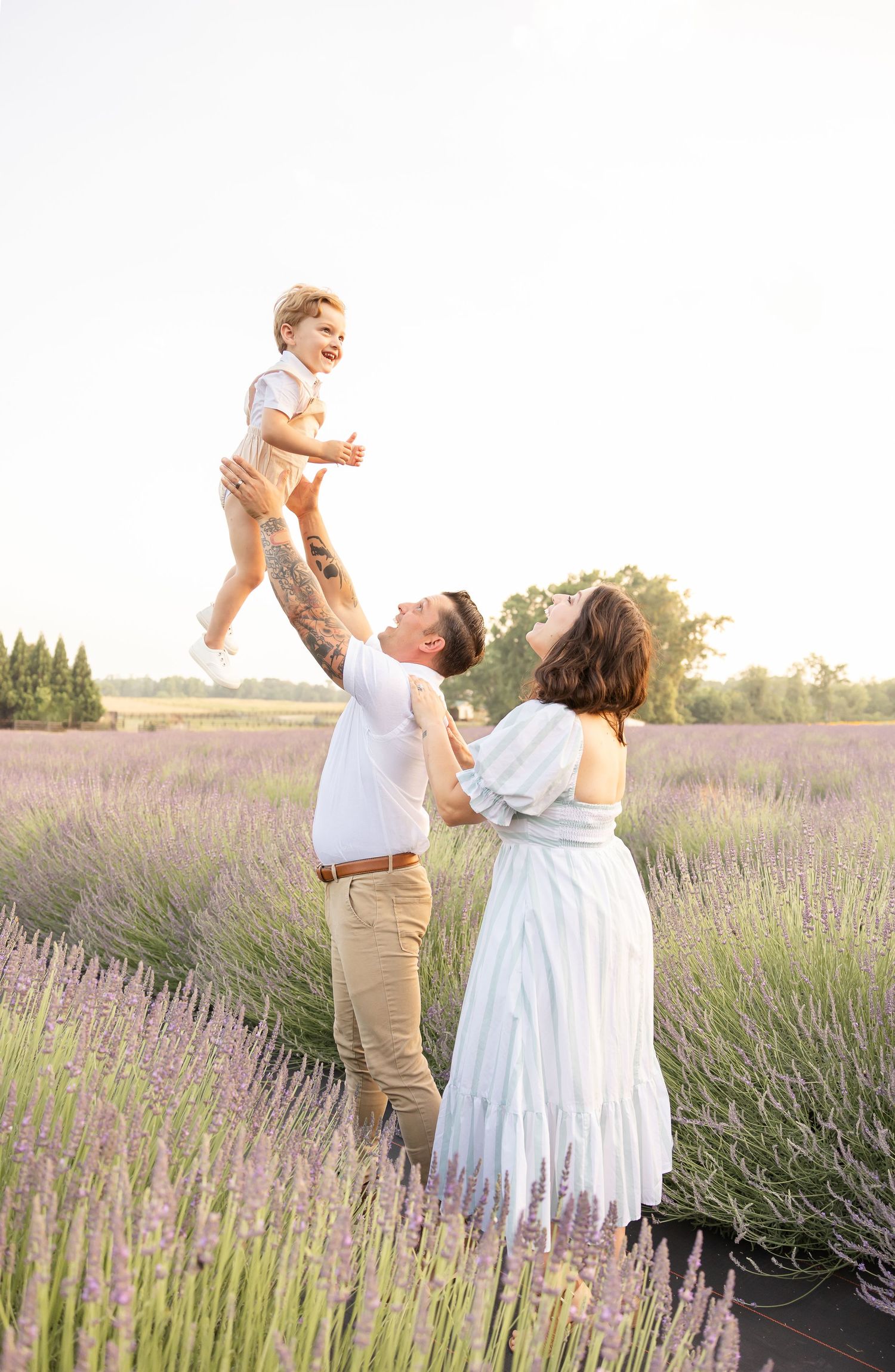 A joyful family in a lavender field, father lifting laughing toddler while mother watches in a white dress.