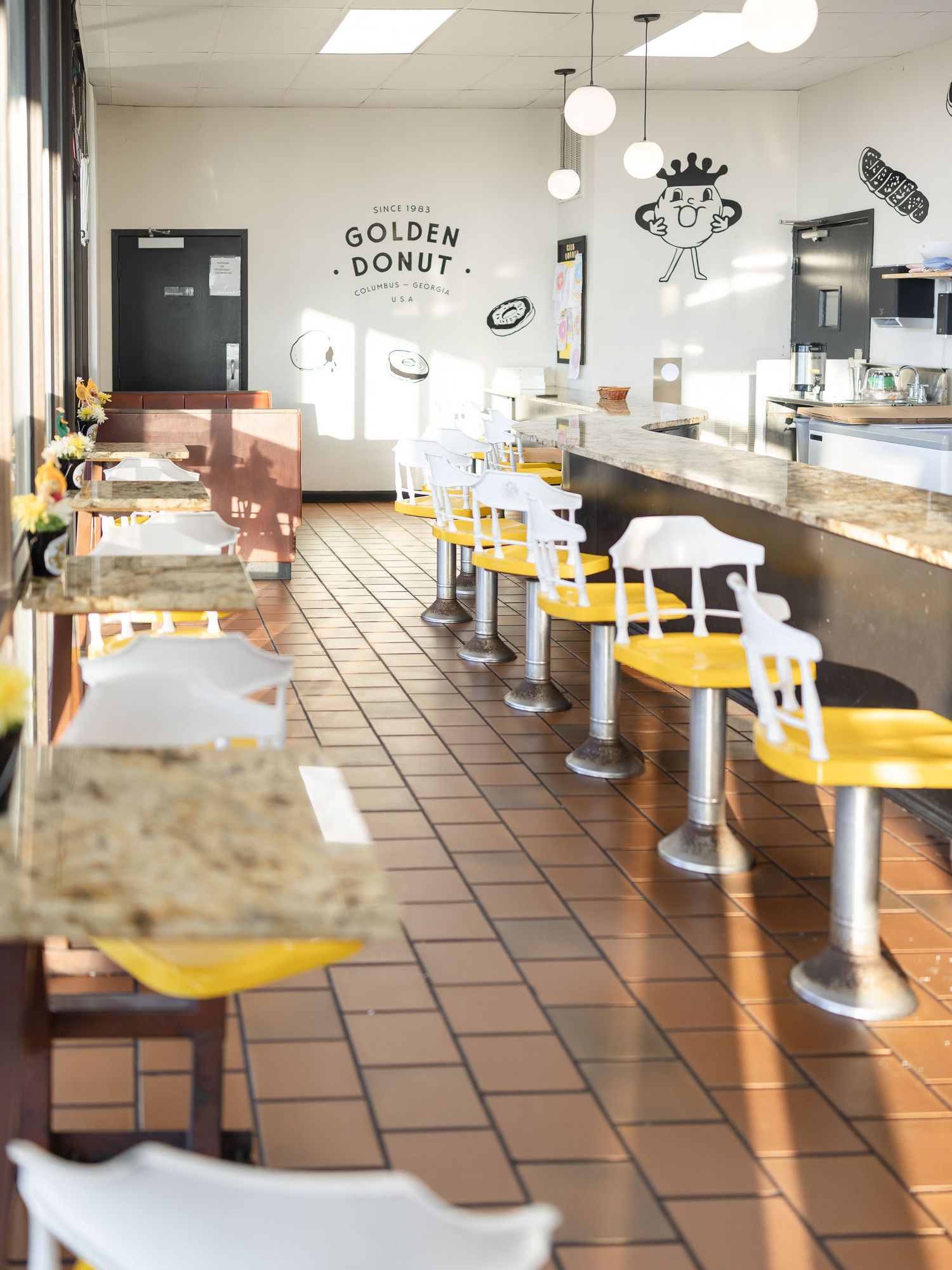 Retro diner interior with yellow counter stools, brown tables, and terracotta tile floors in warm lighting.