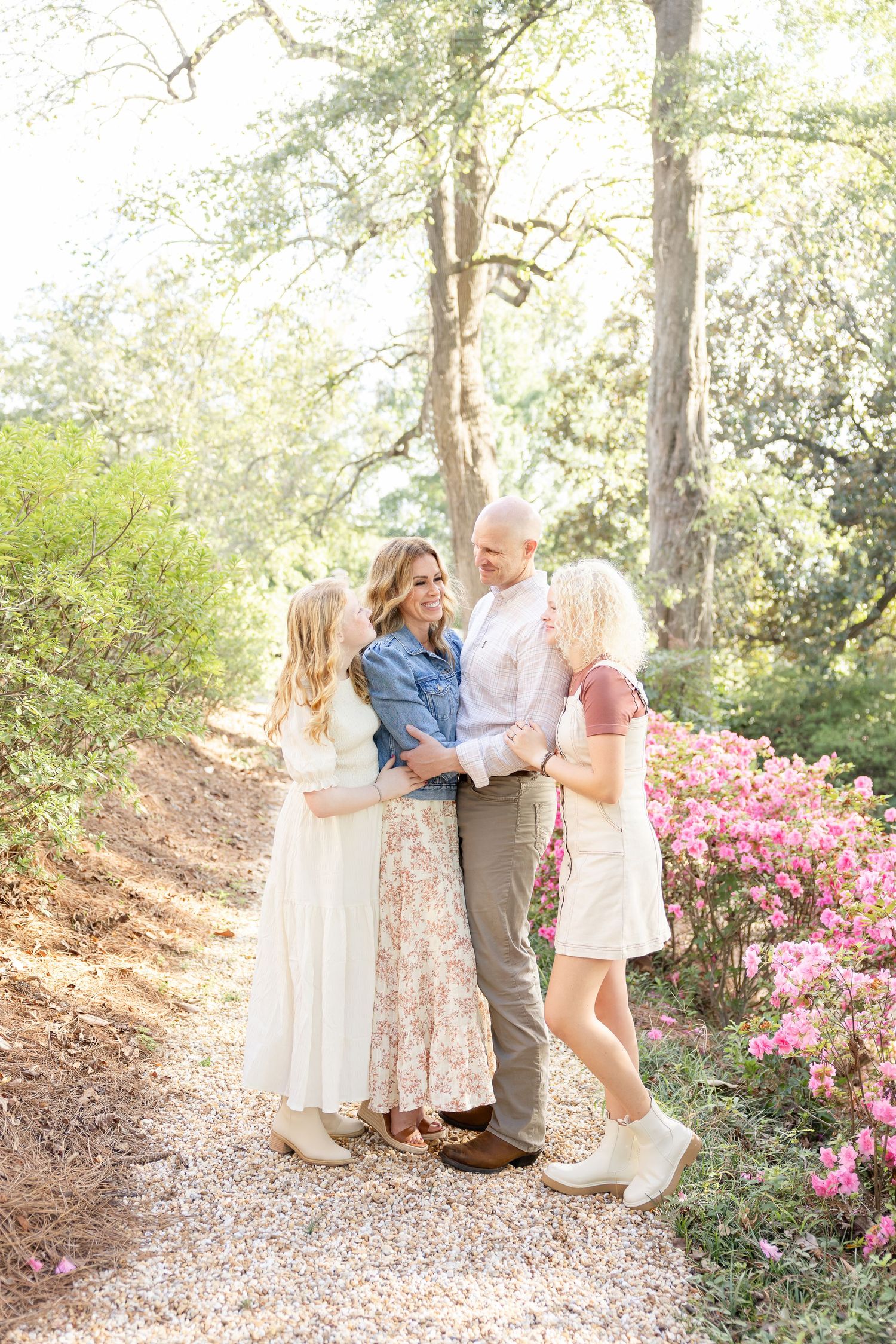 A family embraces on a sunlit path surrounded by blooming pink azaleas and towering trees in a garden setting.