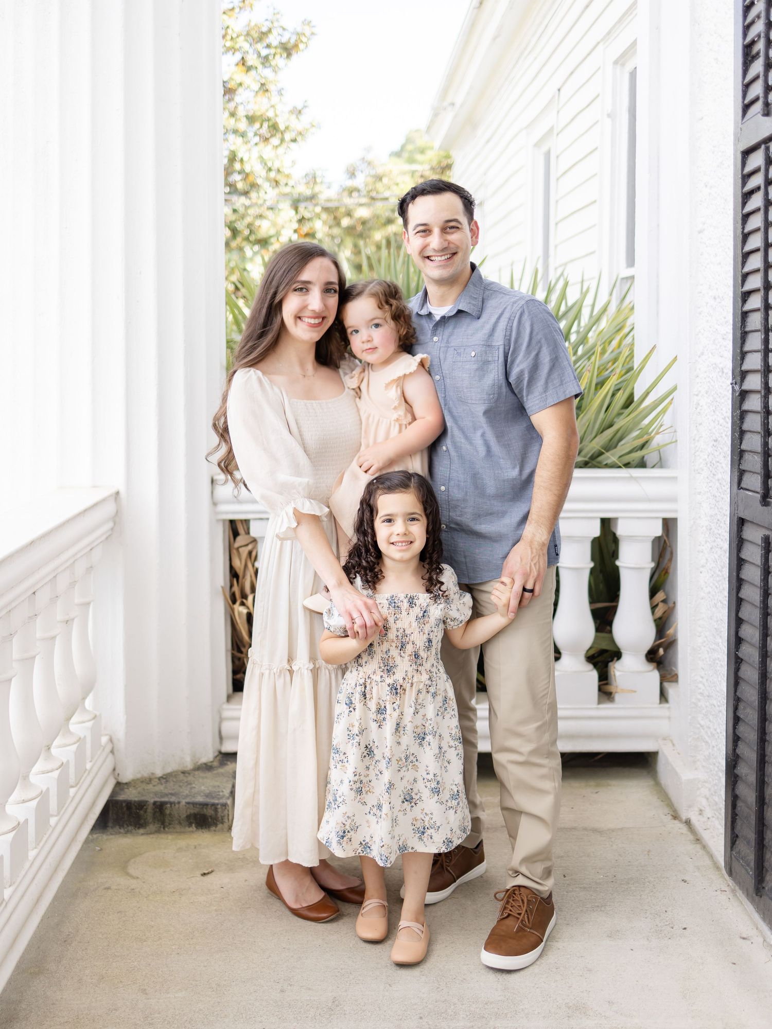 Family of four standing on a white columned porch wearing coordinated neutral-toned outfits for a portrait.