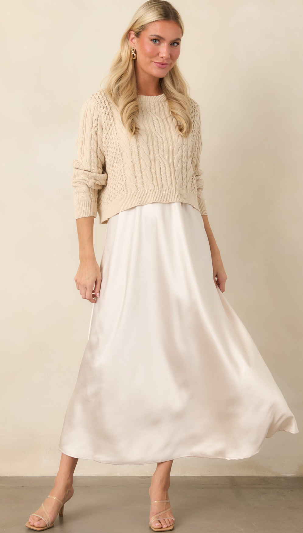 Woman wearing a beige cable-knit cropped sweater with a white satin midi skirt and nude heeled sandals.