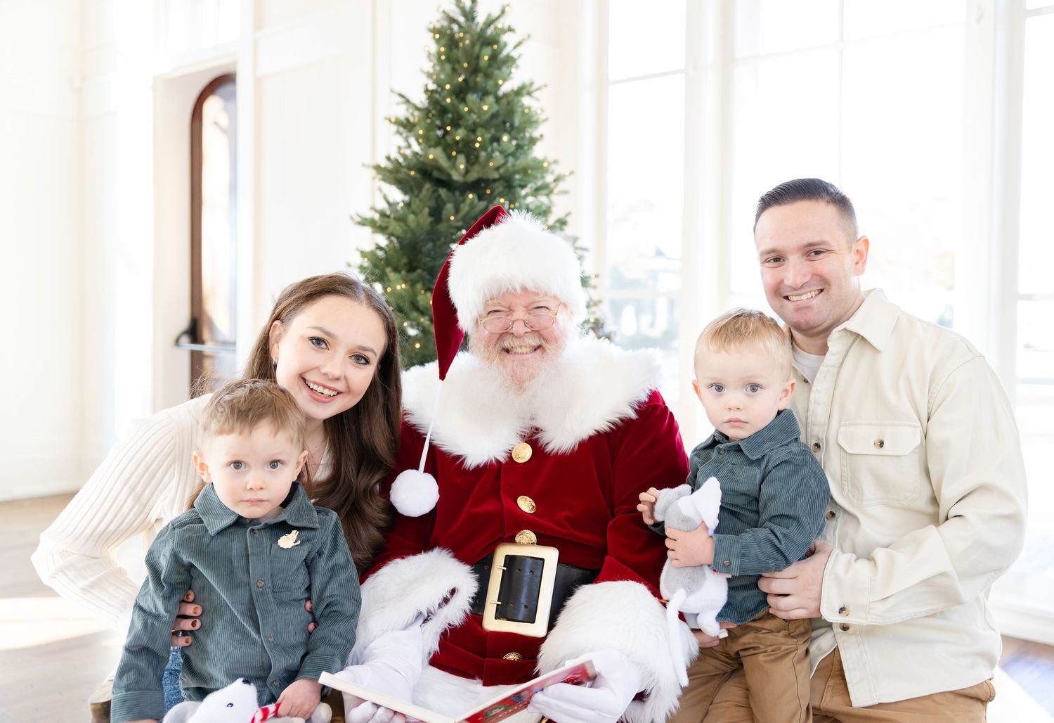 Family posing with Santa Claus in front of Christmas tree, including two young children, holiday portrait.