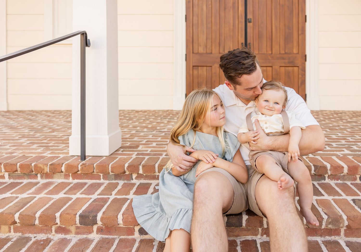 A dad sits on brick steps holding his 2 children during their Columbus, GA summer family portraits.