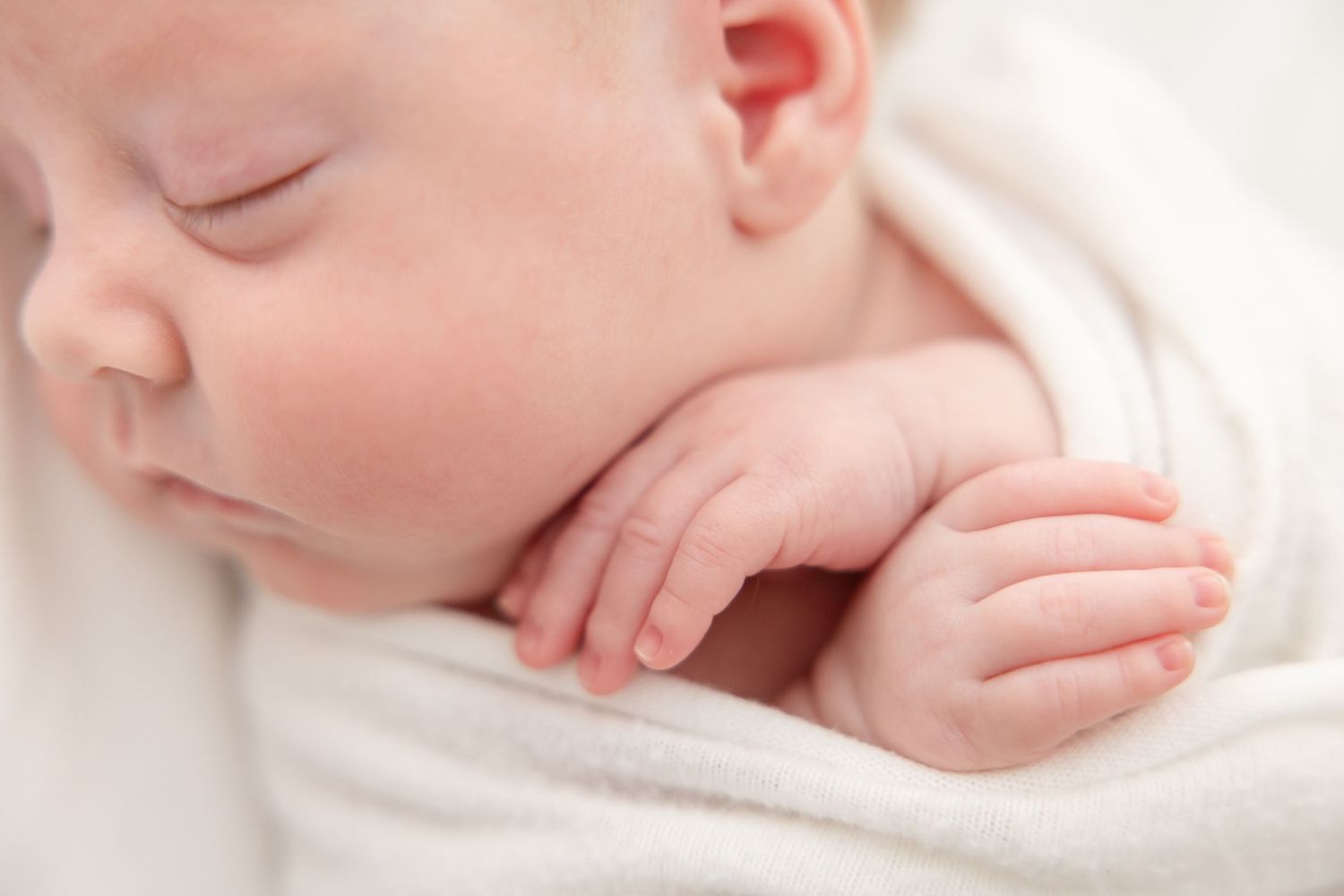A sleeping newborn baby wrapped in soft white fabric with tiny curled fingers visible.