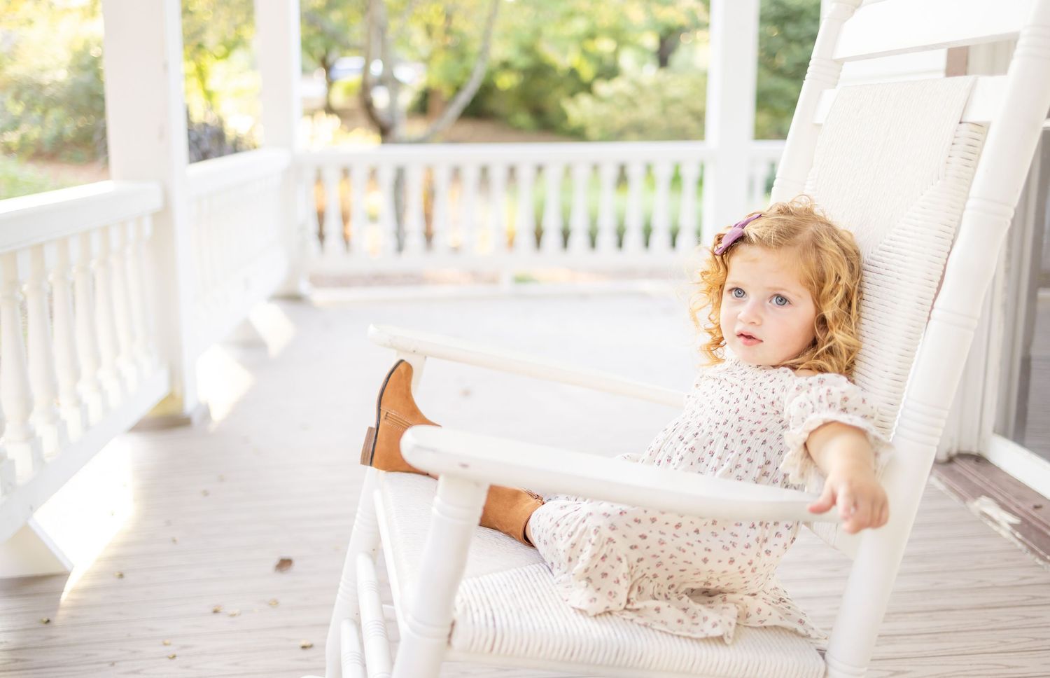 A child in a white floral dress sits back in a rocking chair on a sunny white porch.