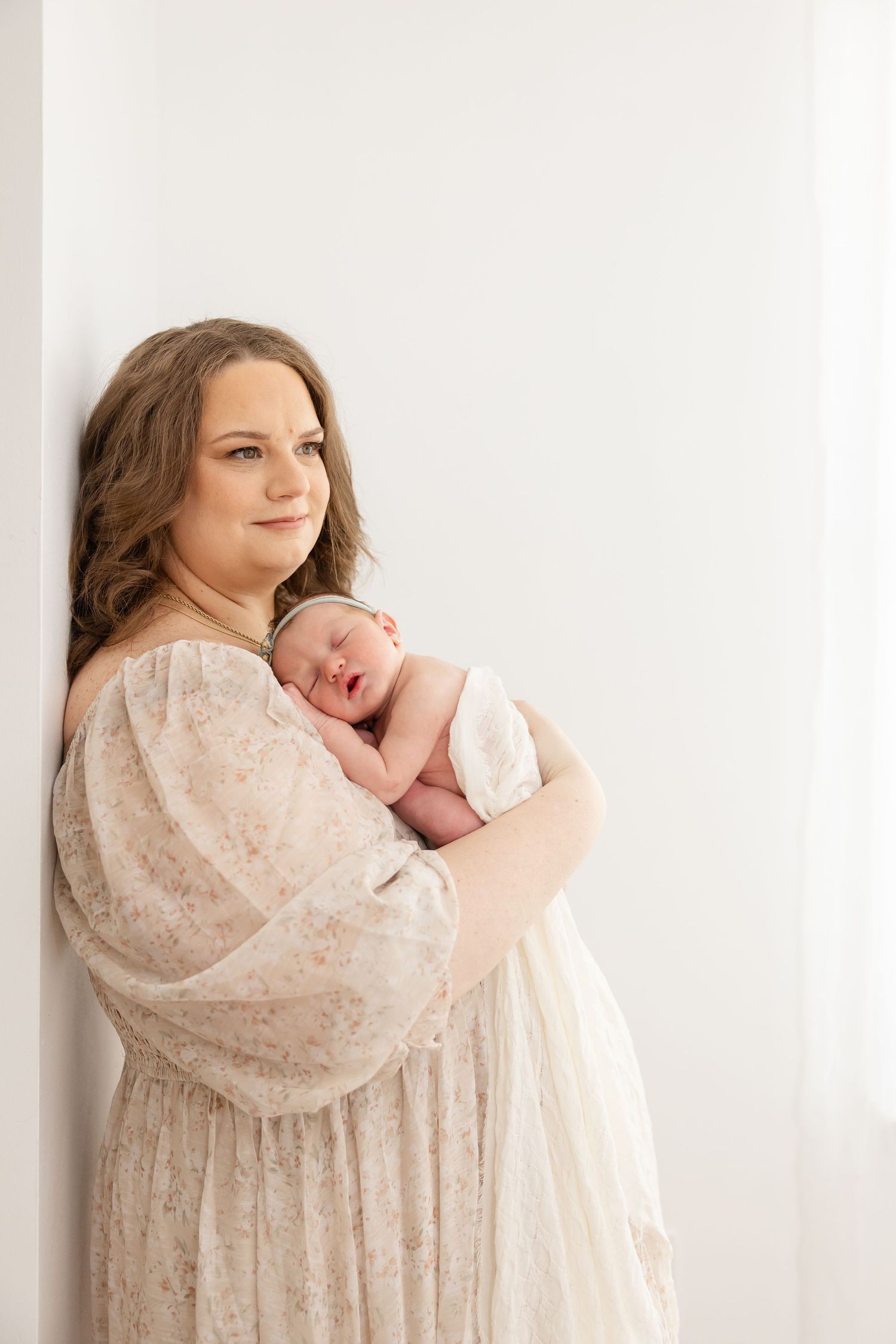 A woman in a flowy cream lace dress holds a sleeping newborn baby while standing against a white wall.
