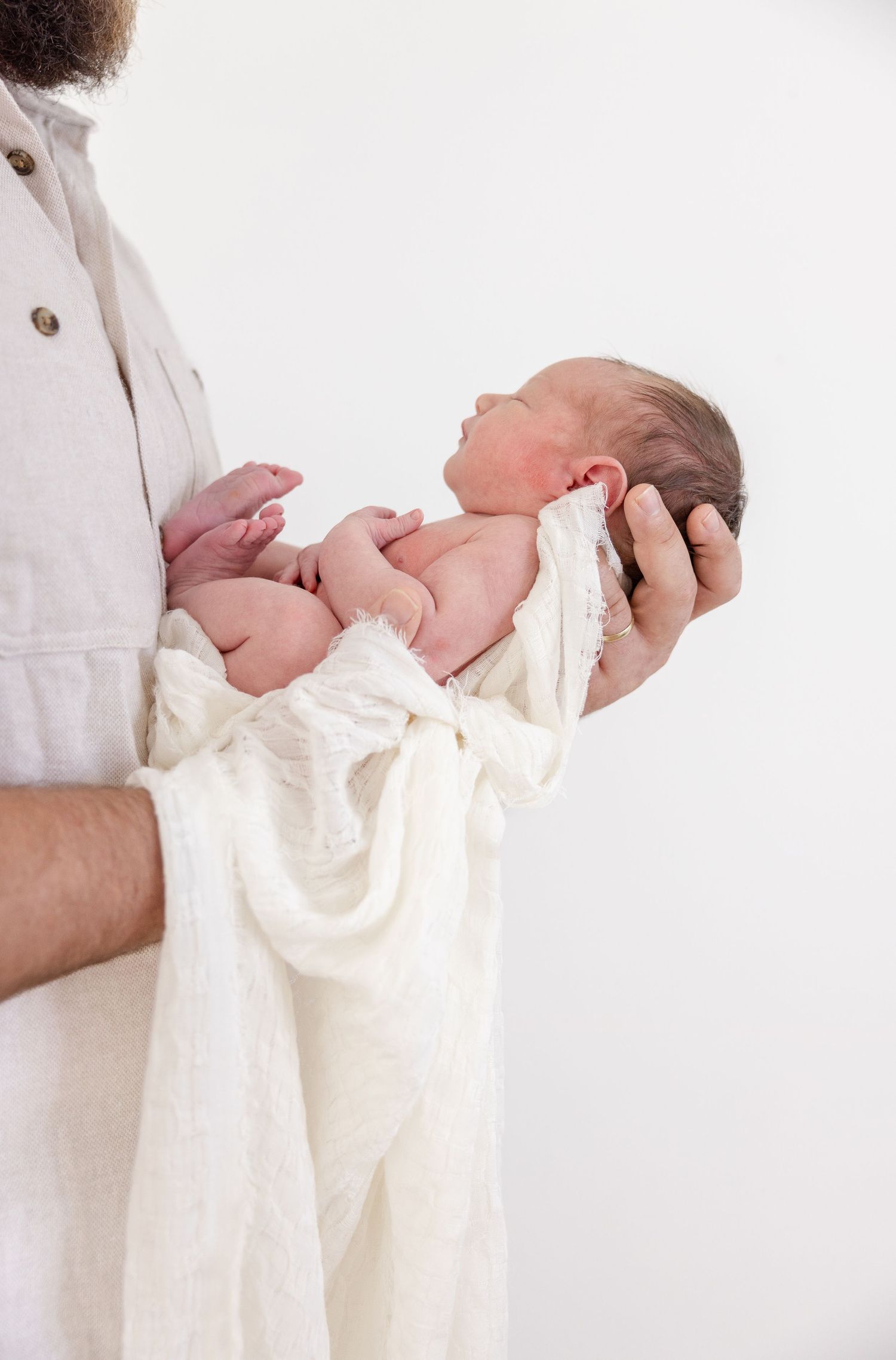 A newborn baby wrapped in a white blanket is being gently cradled in caring arms against a light background.