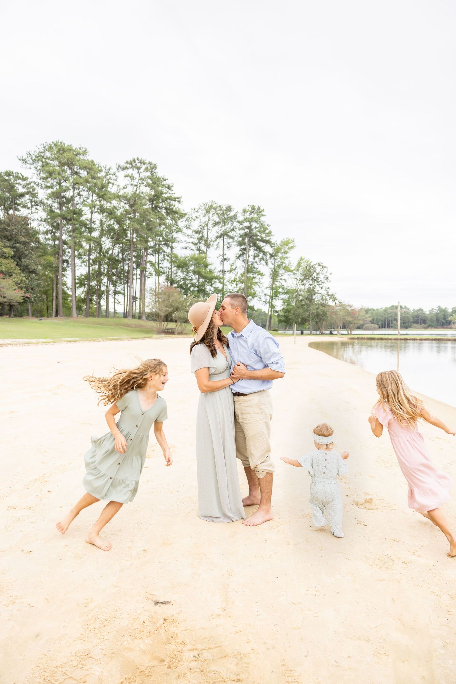 A loving family plays together on a sunlit beach with pine trees and a lake in the background.
