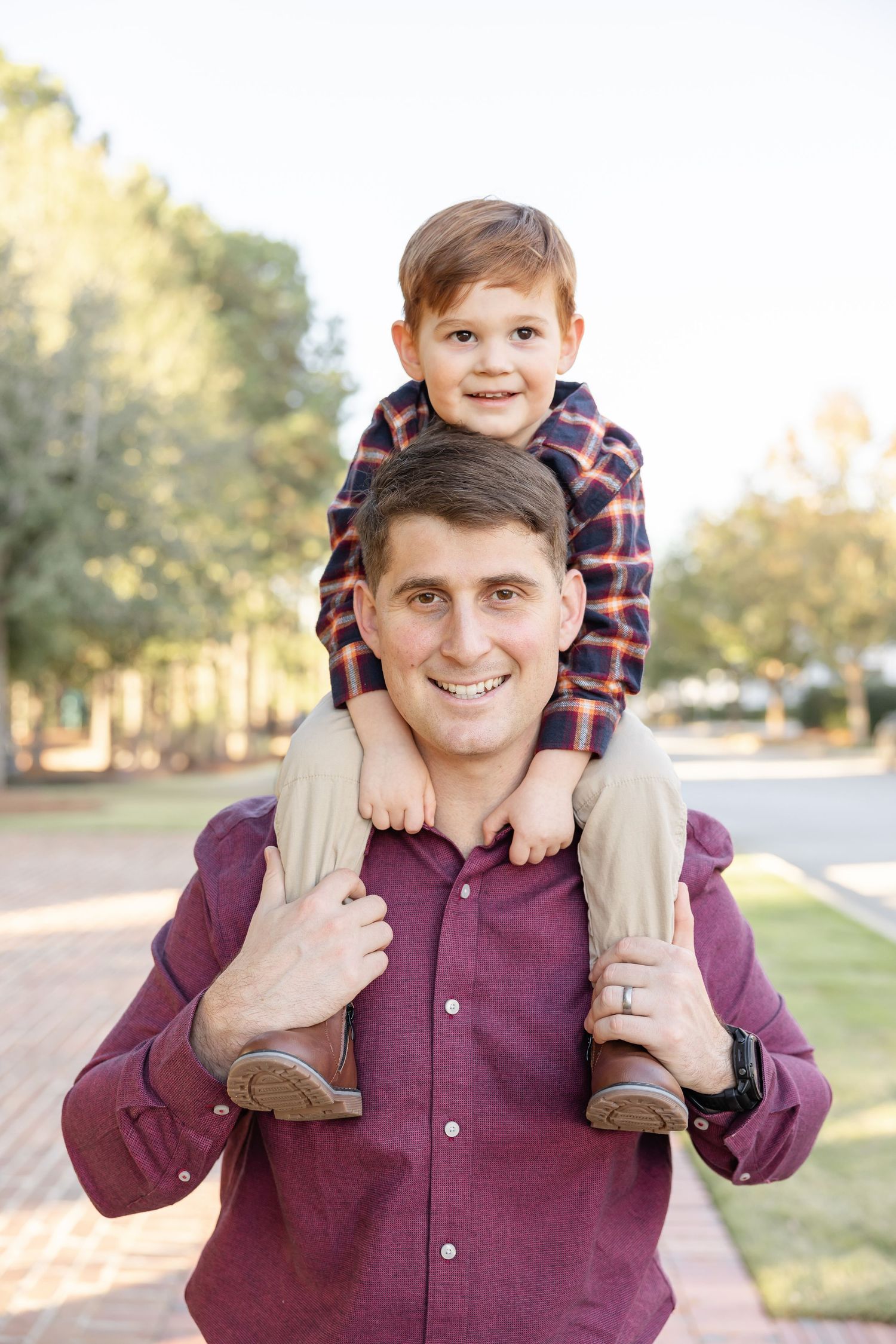 A father in a burgundy button down holds his son on his shoulders as they both smile at the camera at their fall family photography session