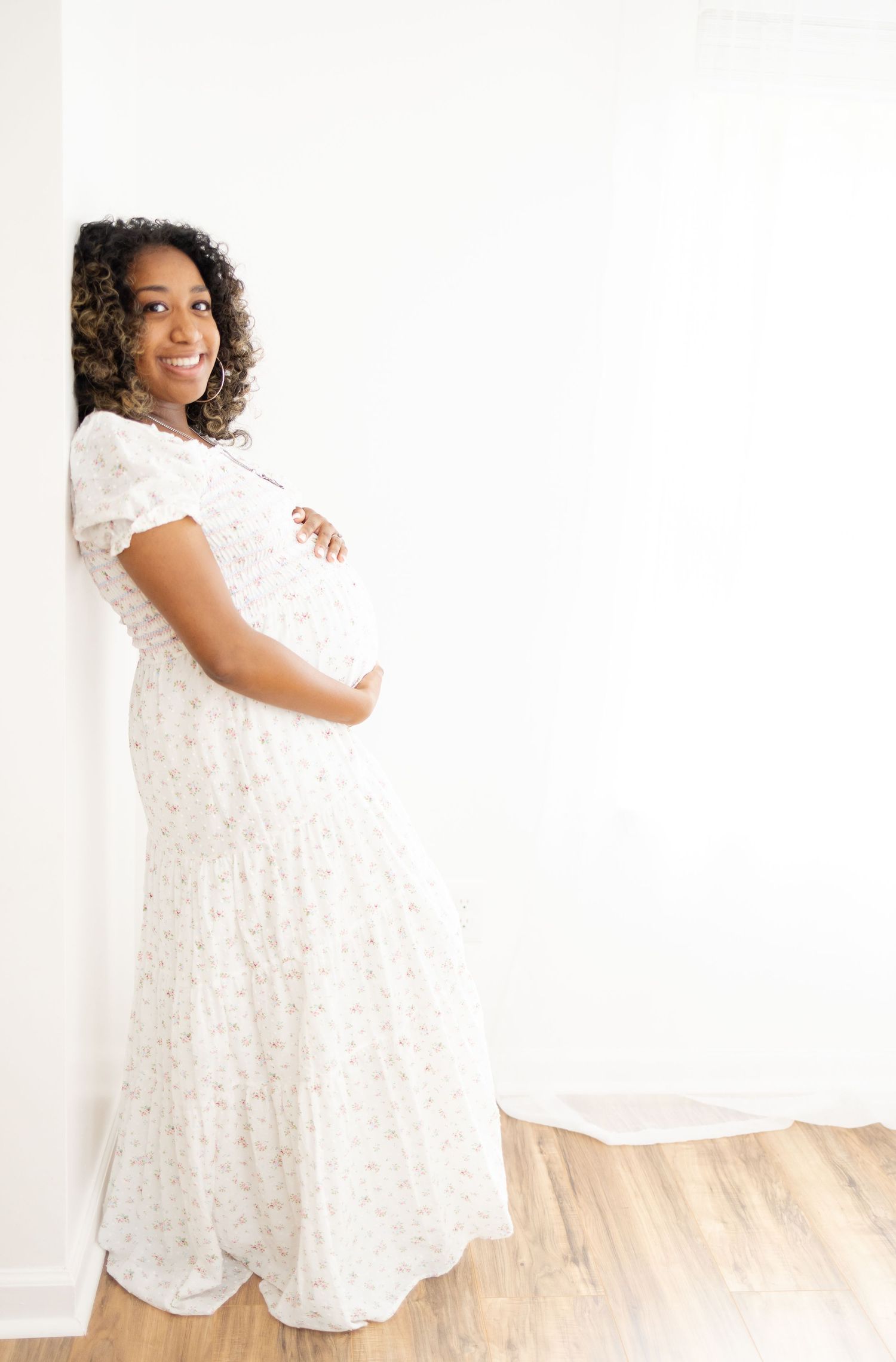 A mother-to-be rests her back against a white wall while holding her belly and smiling at her Columbus, GA Maternity photographer.
