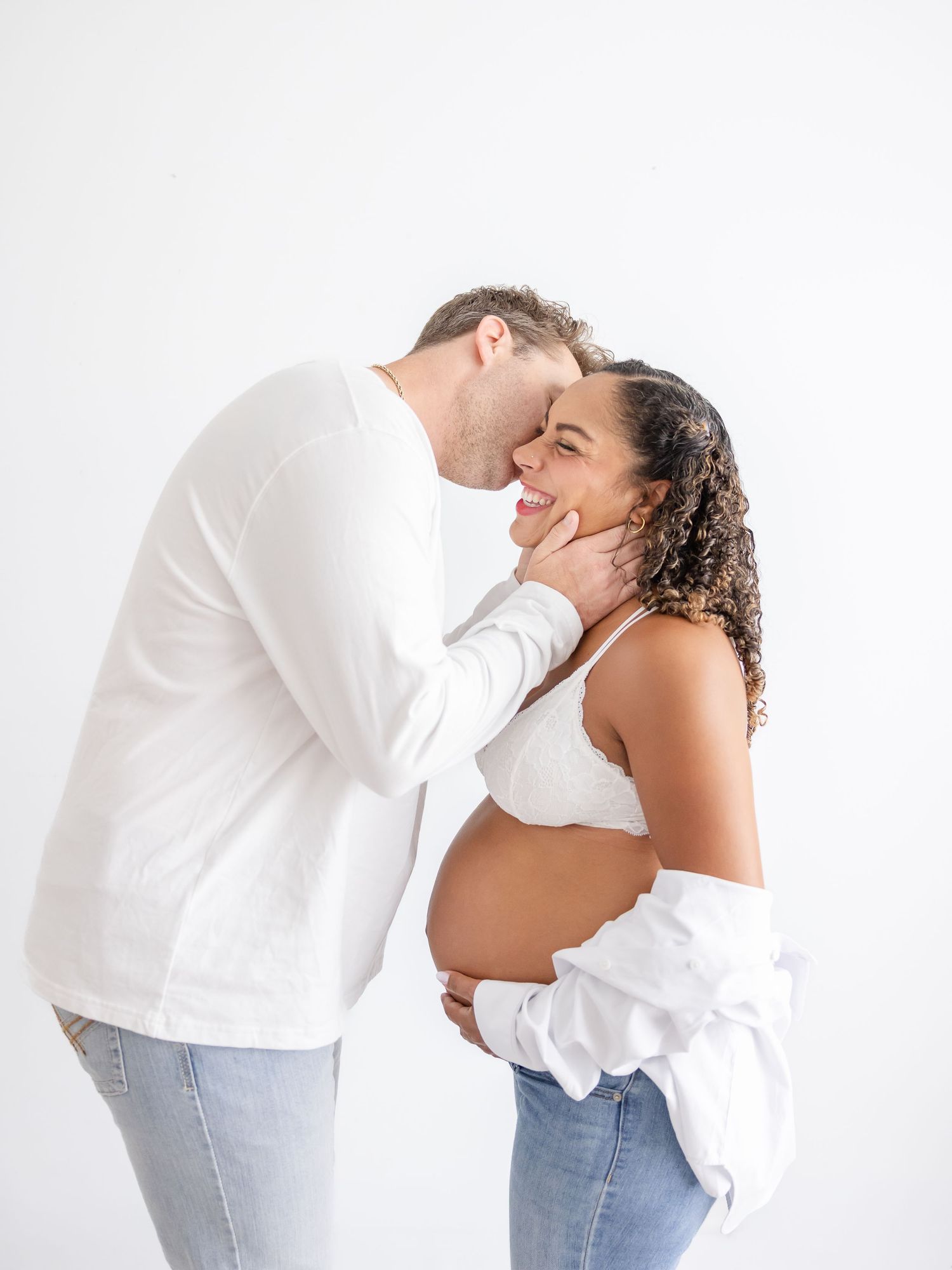 Couple in white outfits sharing a tender moment during maternity photo shoot against white background.