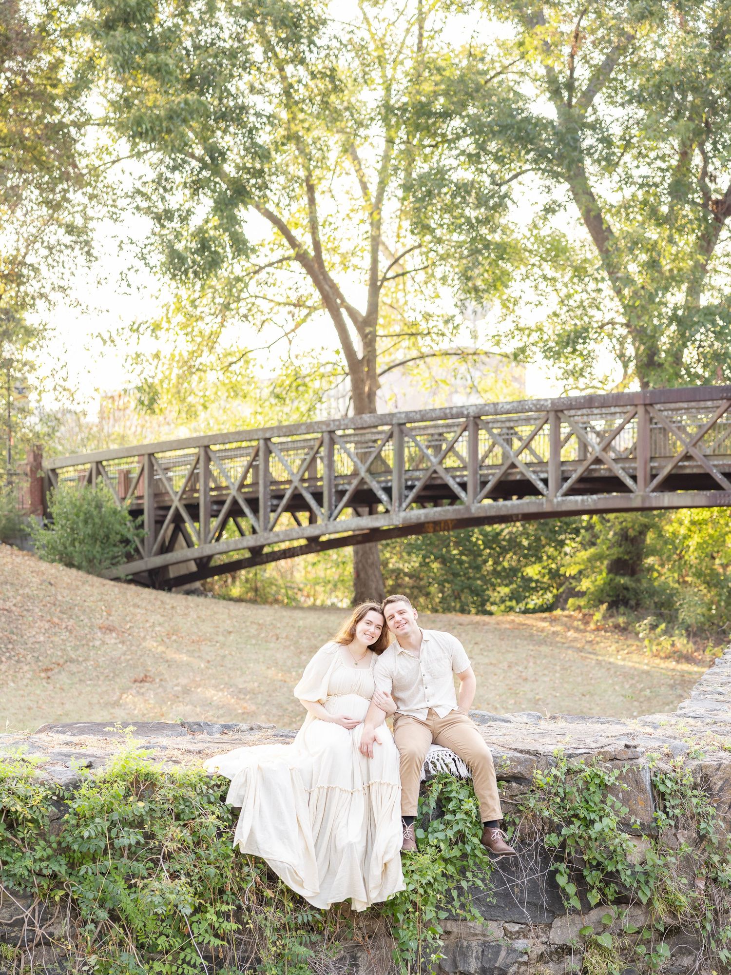 Couple sits on stone wall near wooden bridge surrounded by lush green trees in a romantic outdoor maternity photo.