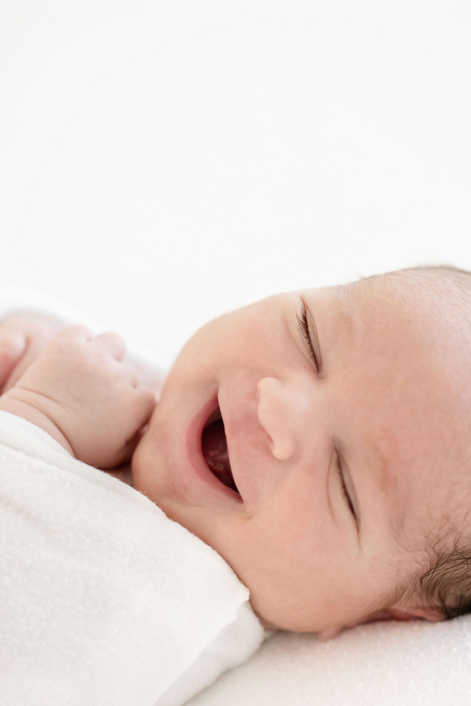 Newborn baby smiling with eyes closed, wrapped in a white blanket, lying on a white surface by Columbus, GA photographer.
