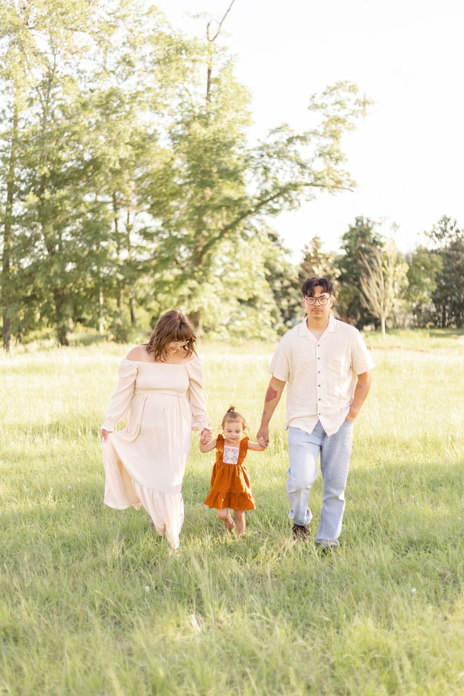 Family of three walking in a sunny green field, parents holding toddler's hands in a warm outdoor photo session.