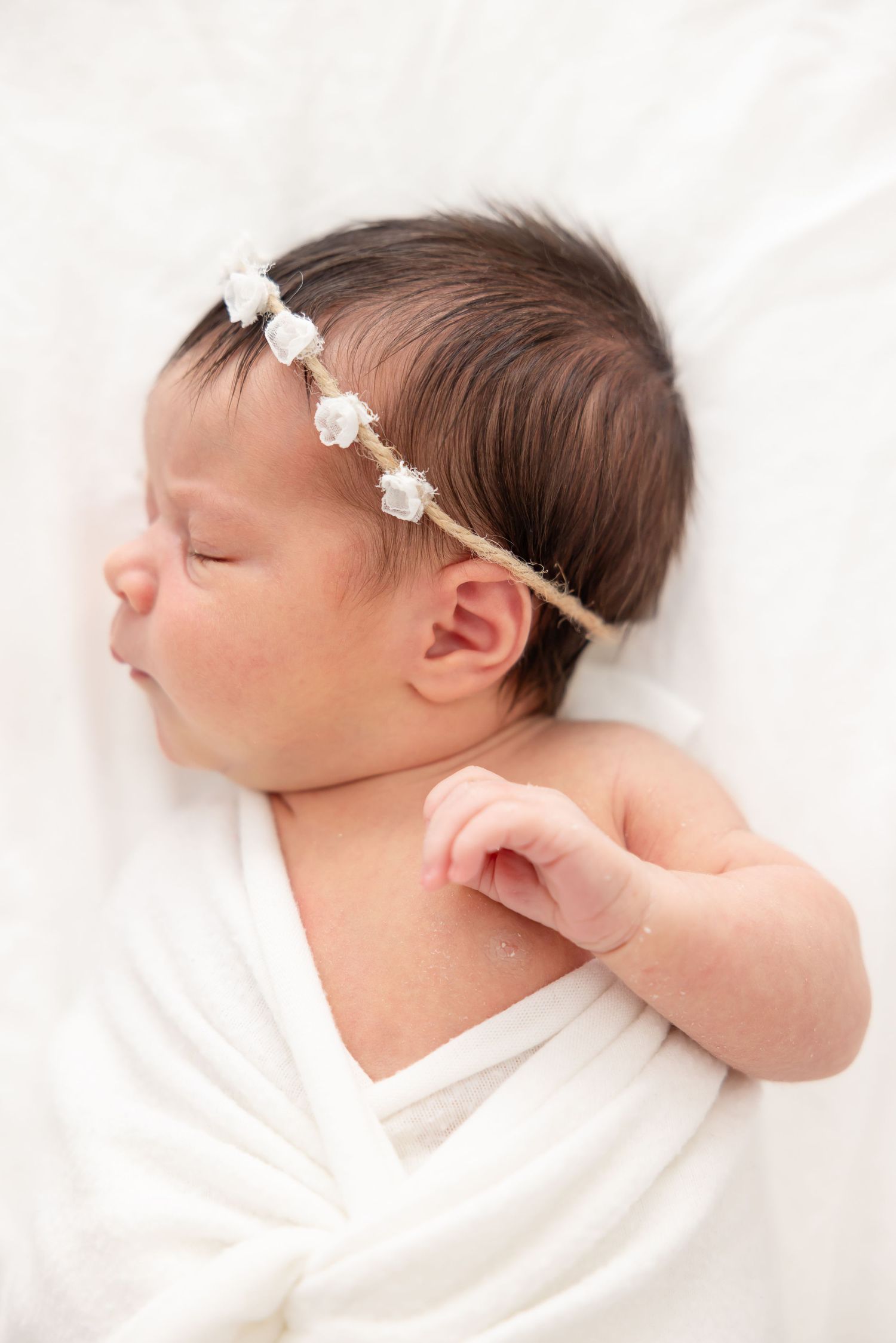 Newborn baby wrapped in white fabric sleeping peacefully on white blanket wearing delicate floral headband.