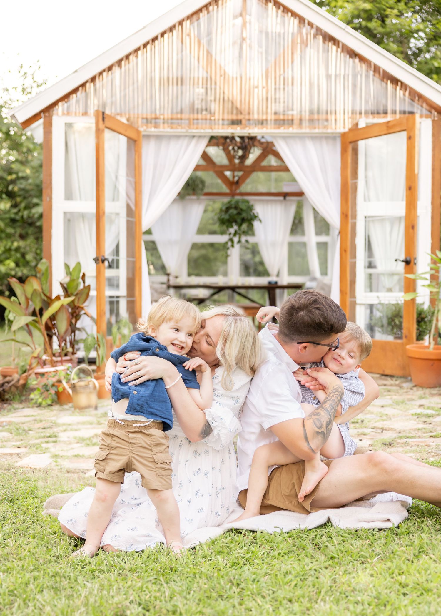 A family shares tender moments together on a blanket in front of a rustic greenhouse with flowing white curtains.