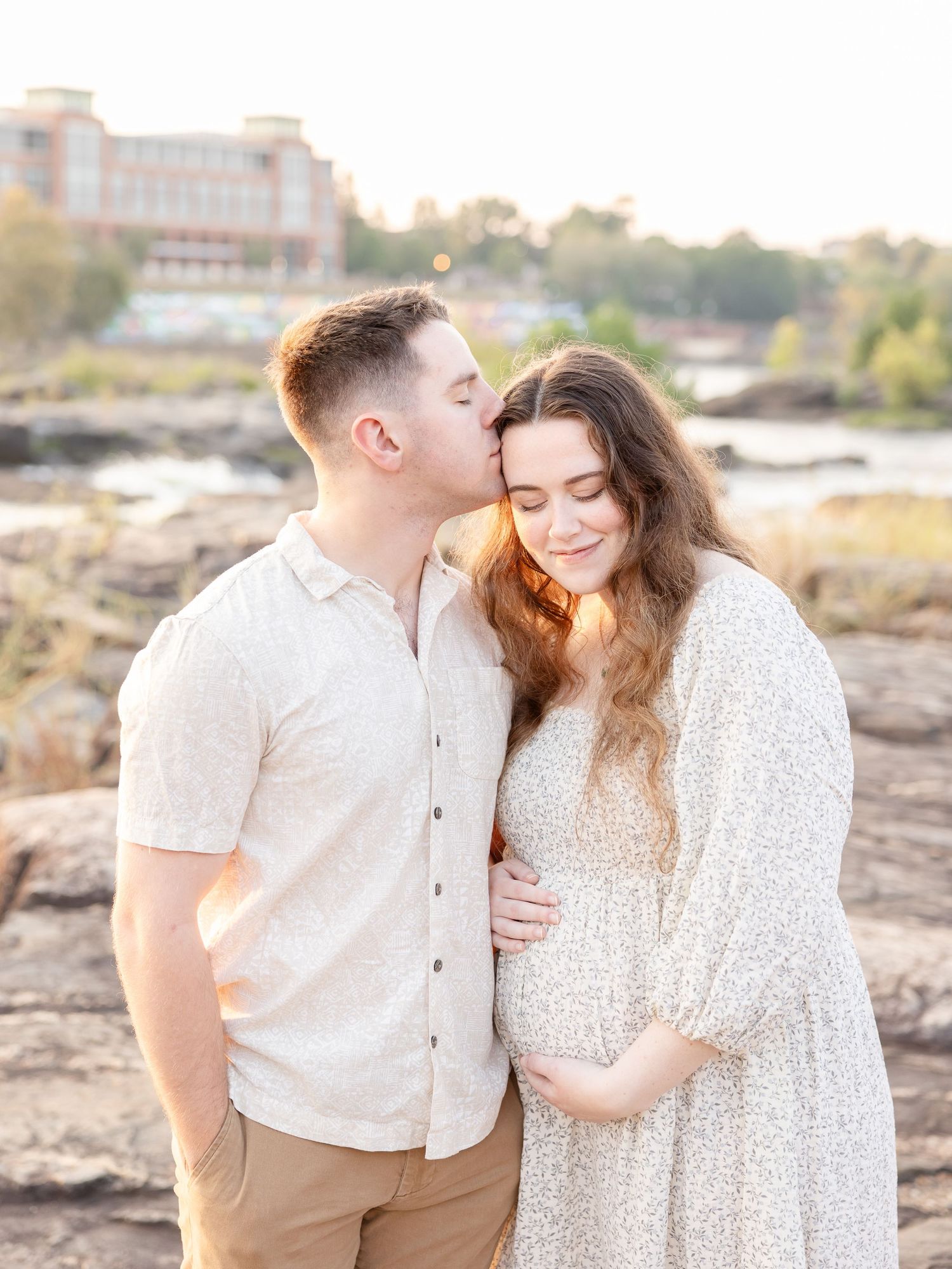 A couple shares a tender moment outdoors at golden hour, the man kissing his pregnant partner's cheek near a river.