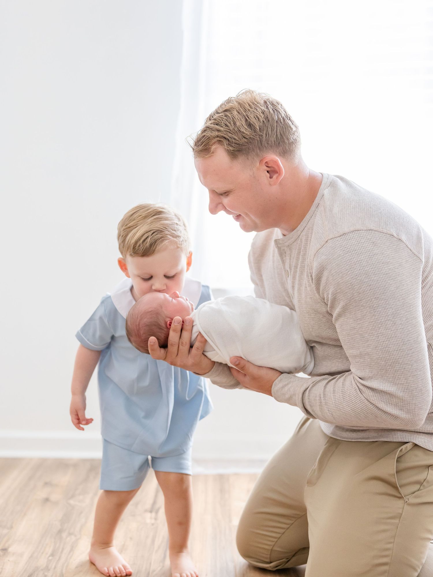 Tender family moment as big brother gives his newborn brother in dad’s arms a kiss in a bright, minimalist room by Columbus, GA photographer