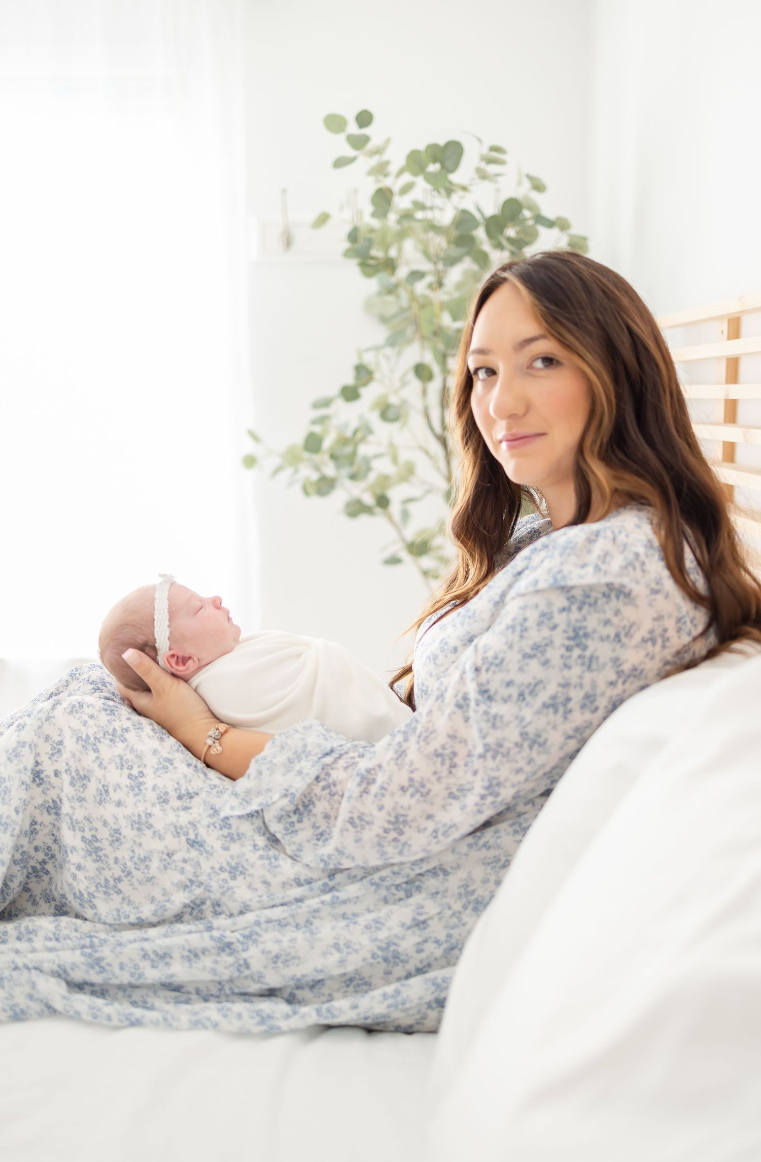 A mother wearing a floral dress relaxes on white bed in a bright, airy bedroom holding her newborn by Columbus, GA newborn photographer.