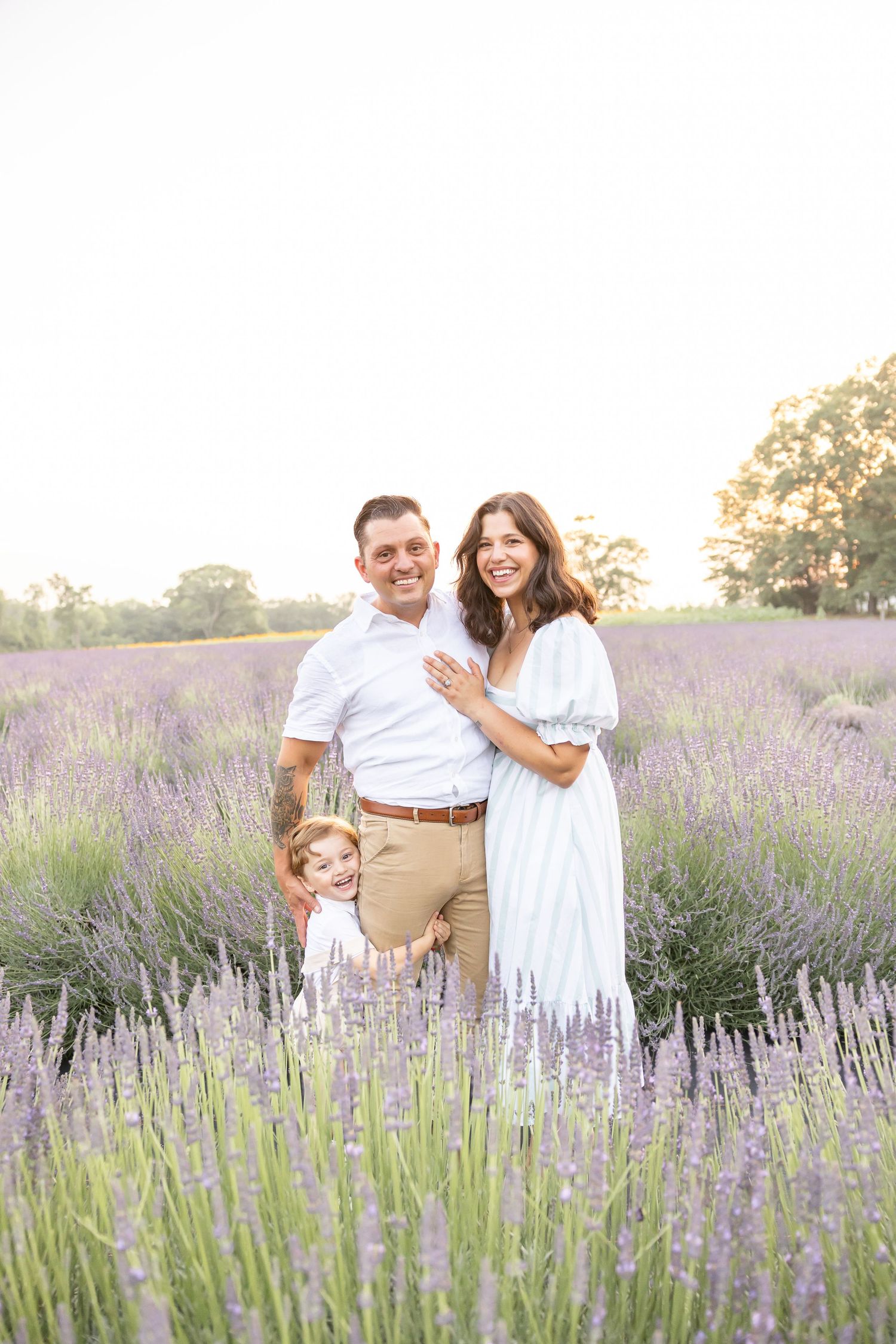 Family of three posing in a blooming lavender field, parents smiling with child, golden hour lighting.