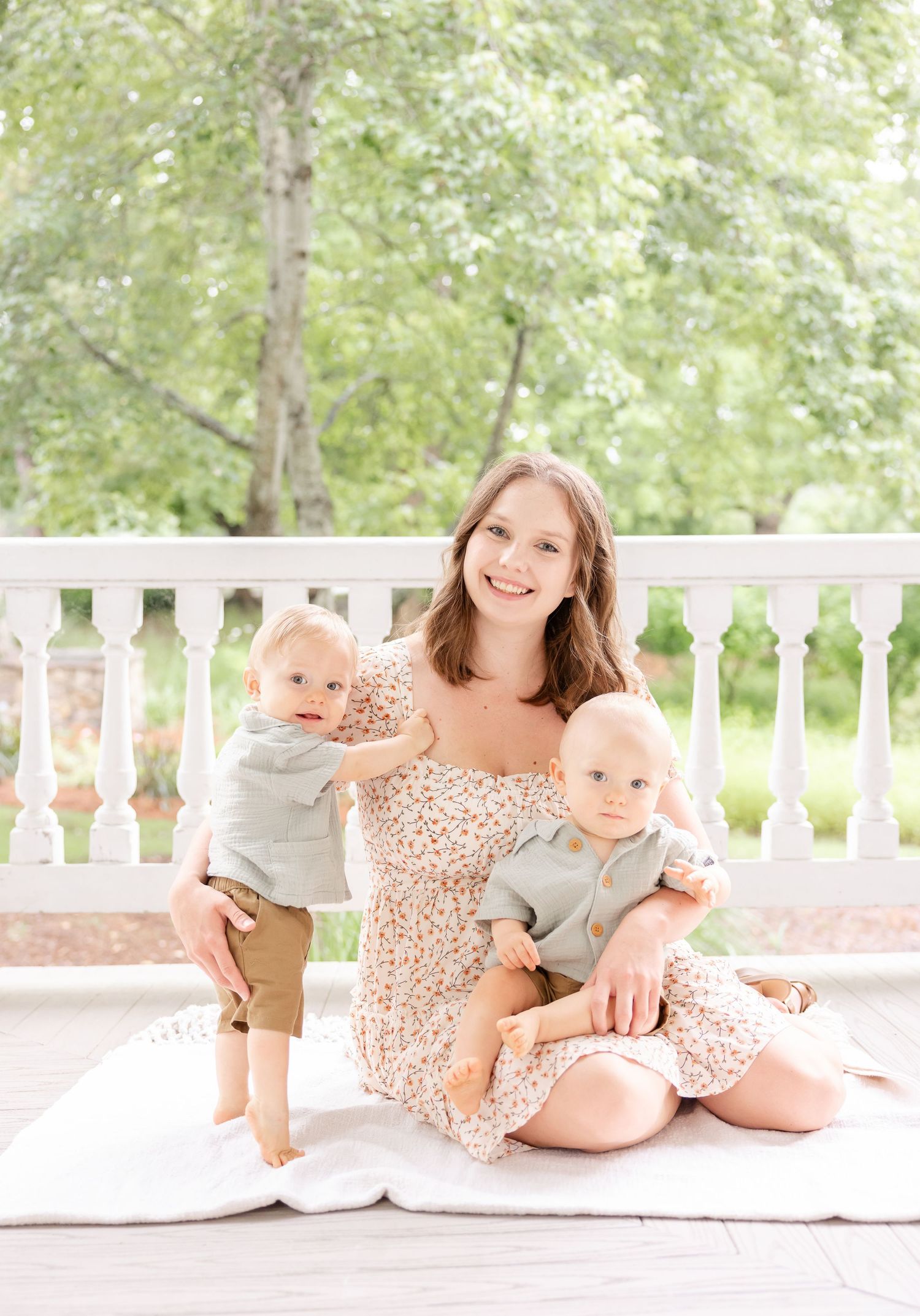 A mother in floral dress sits on a white porch with trees in the background holding her twin boys at their Columbus, GA milestone portraits.