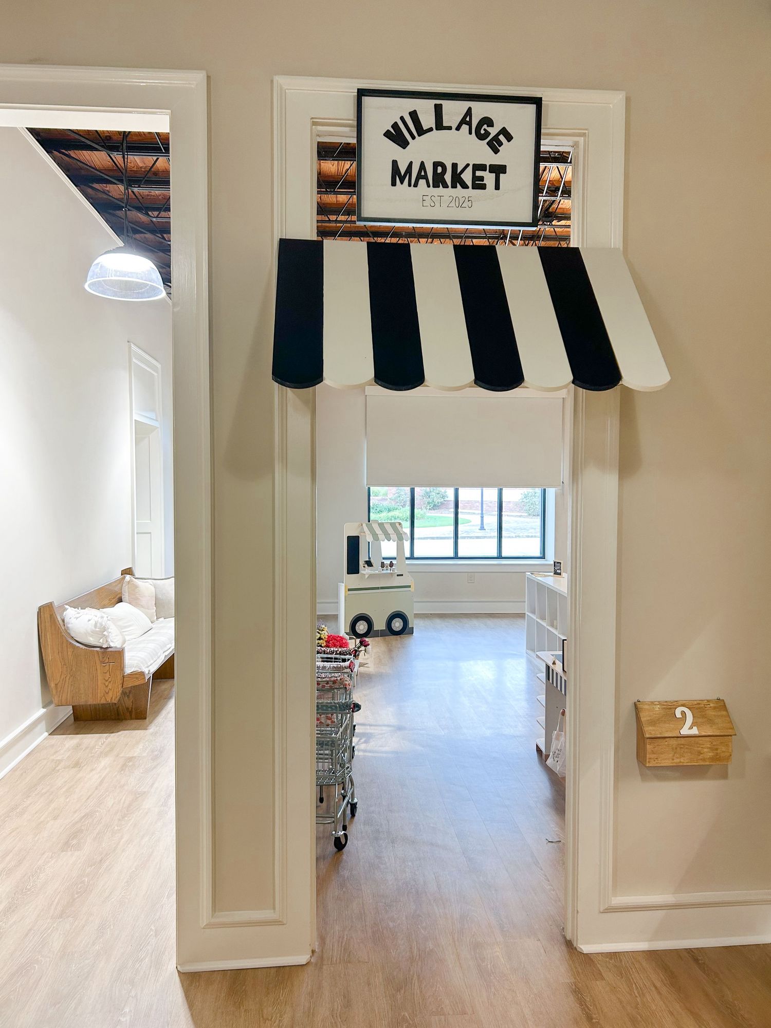 A playful children's play area with a black and white 'Village Market' sign and striped awning mounted on white walls.