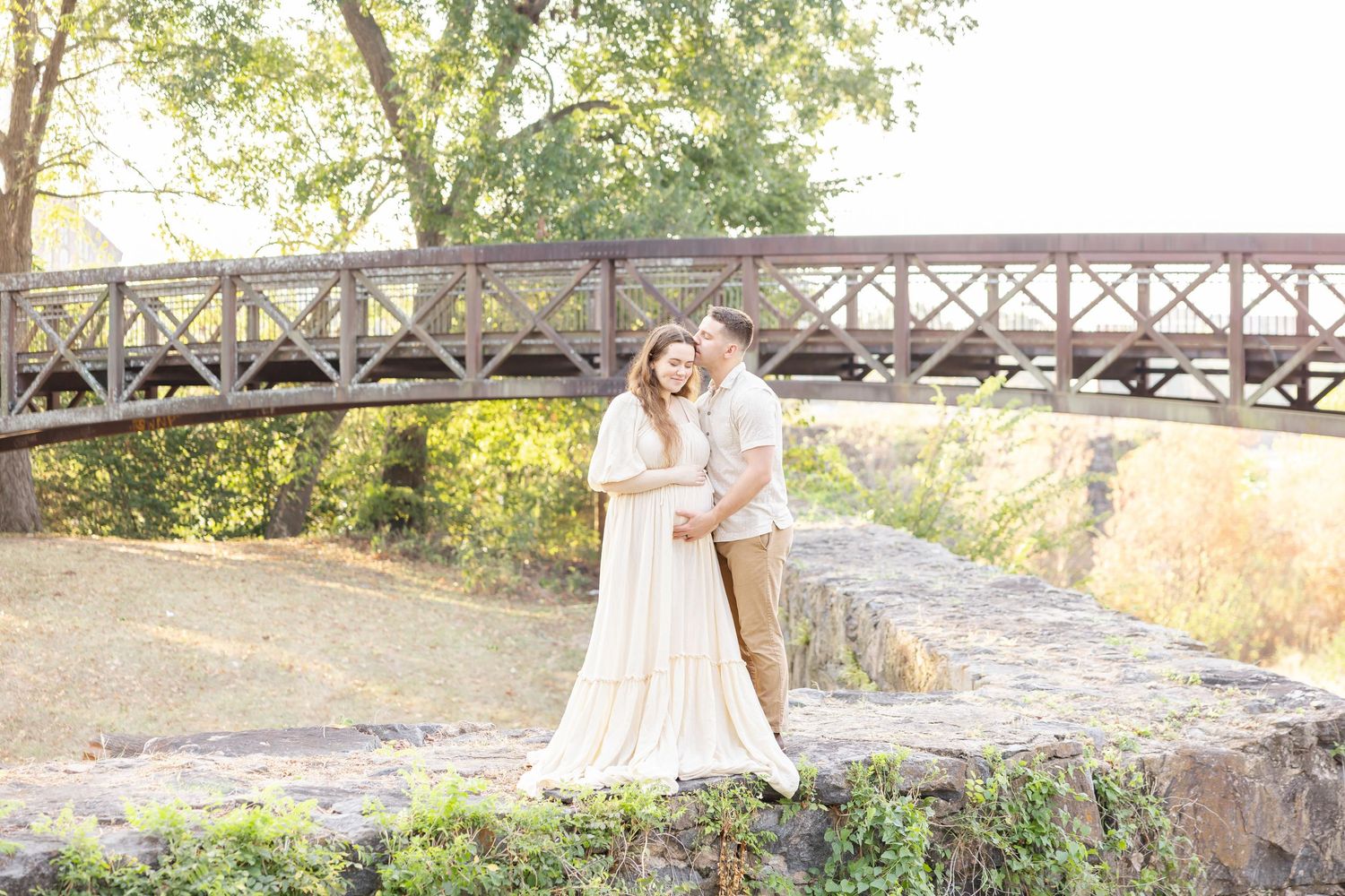 Couple embracing on rocky ledge with wooden bridge and lush trees in soft natural light, woman in flowing white gown.