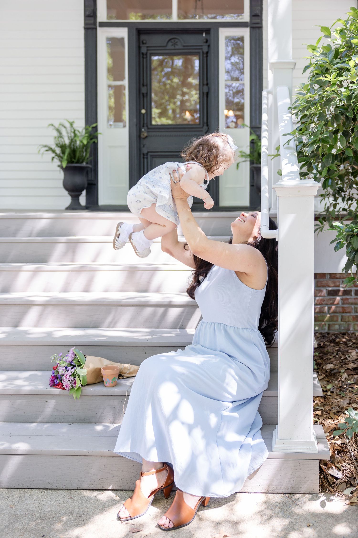 A mother in a blue dress lifts her toddler on sunny porch steps, both smiling joyfully.