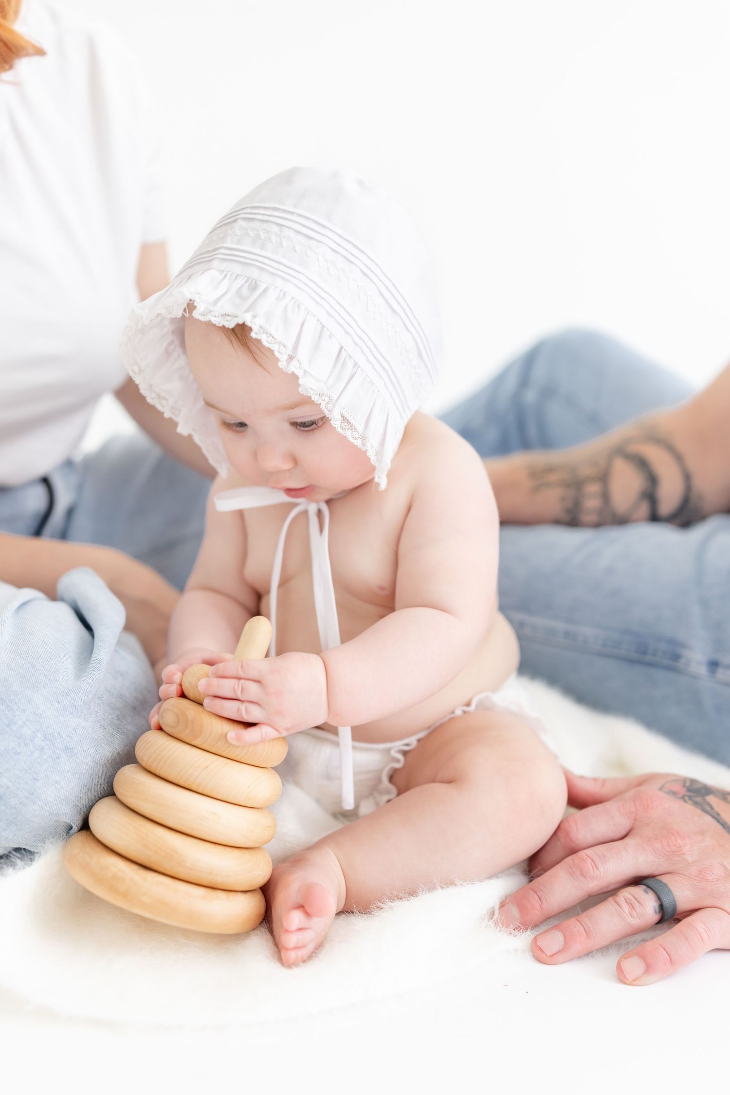 Baby in white bonnet plays with wooden stacking rings toy while parents sit nearby on white surface.