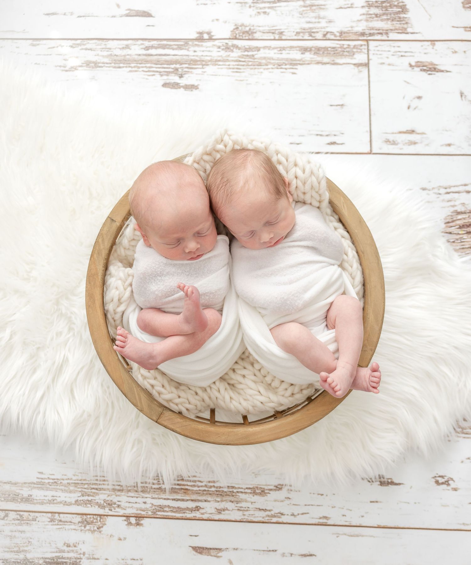 Two newborn twins sleeping peacefully in a wooden bowl with chunky knit blanket on white fur rug.