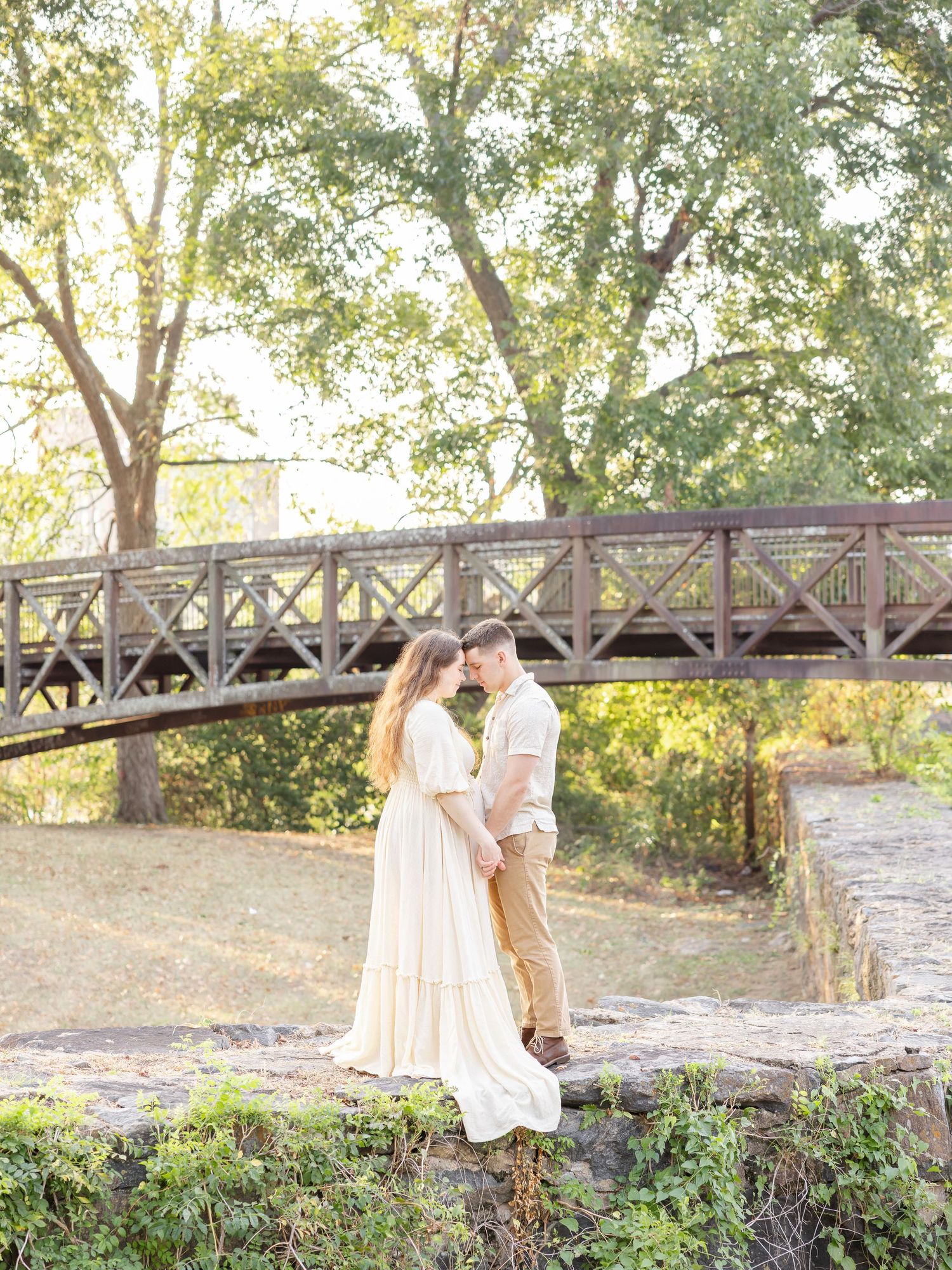 Couple shares a romantic moment on a rustic rock wall surrounded by lush greenery in golden sunlight by Columbus, GA maternity photographer.