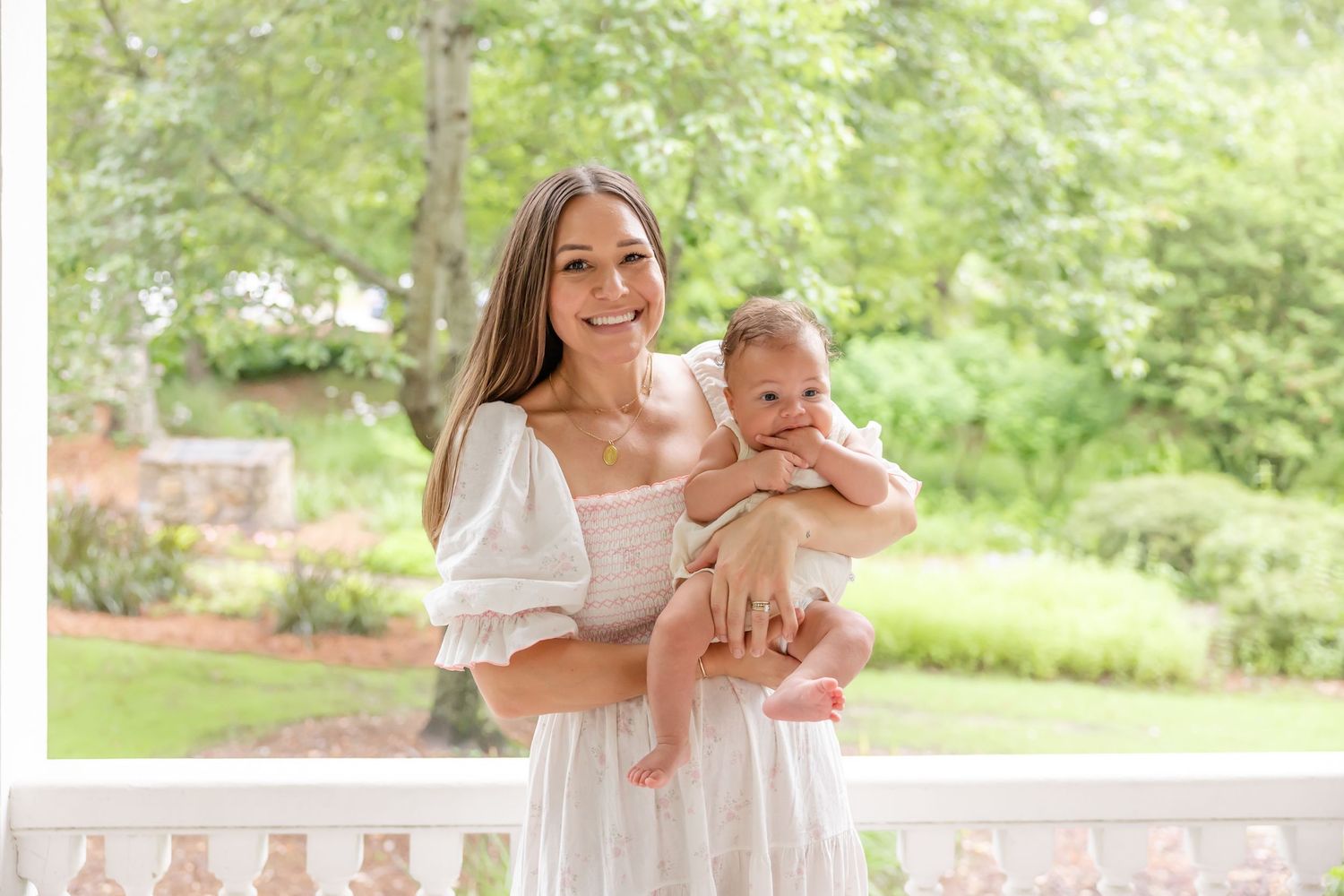 A mom and her newborn on a white porch with greenery and natural lighting in the background by Columbus, GA family photographer.